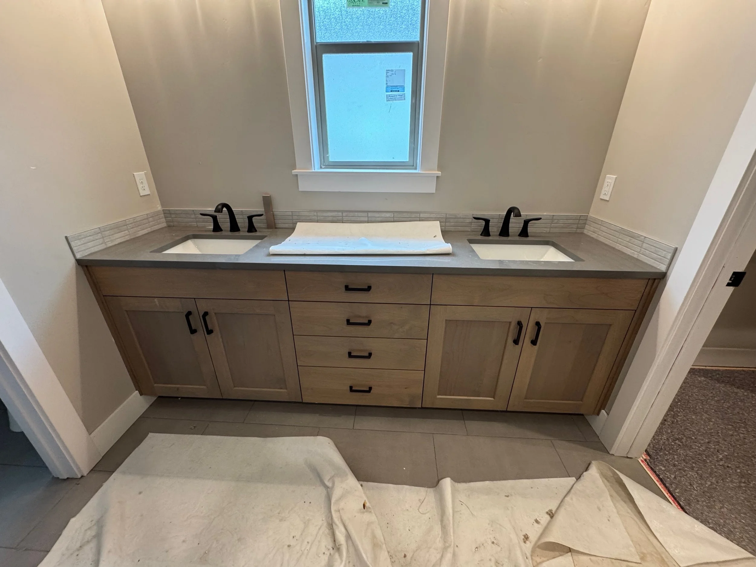 Bathroom vanity with a dual sink countertop, black faucets, light wood cabinets, and a window above. A white towel is draped over the counter and a white rug is placed in front of the vanity. There are electrical outlets on both sides of the countertop.