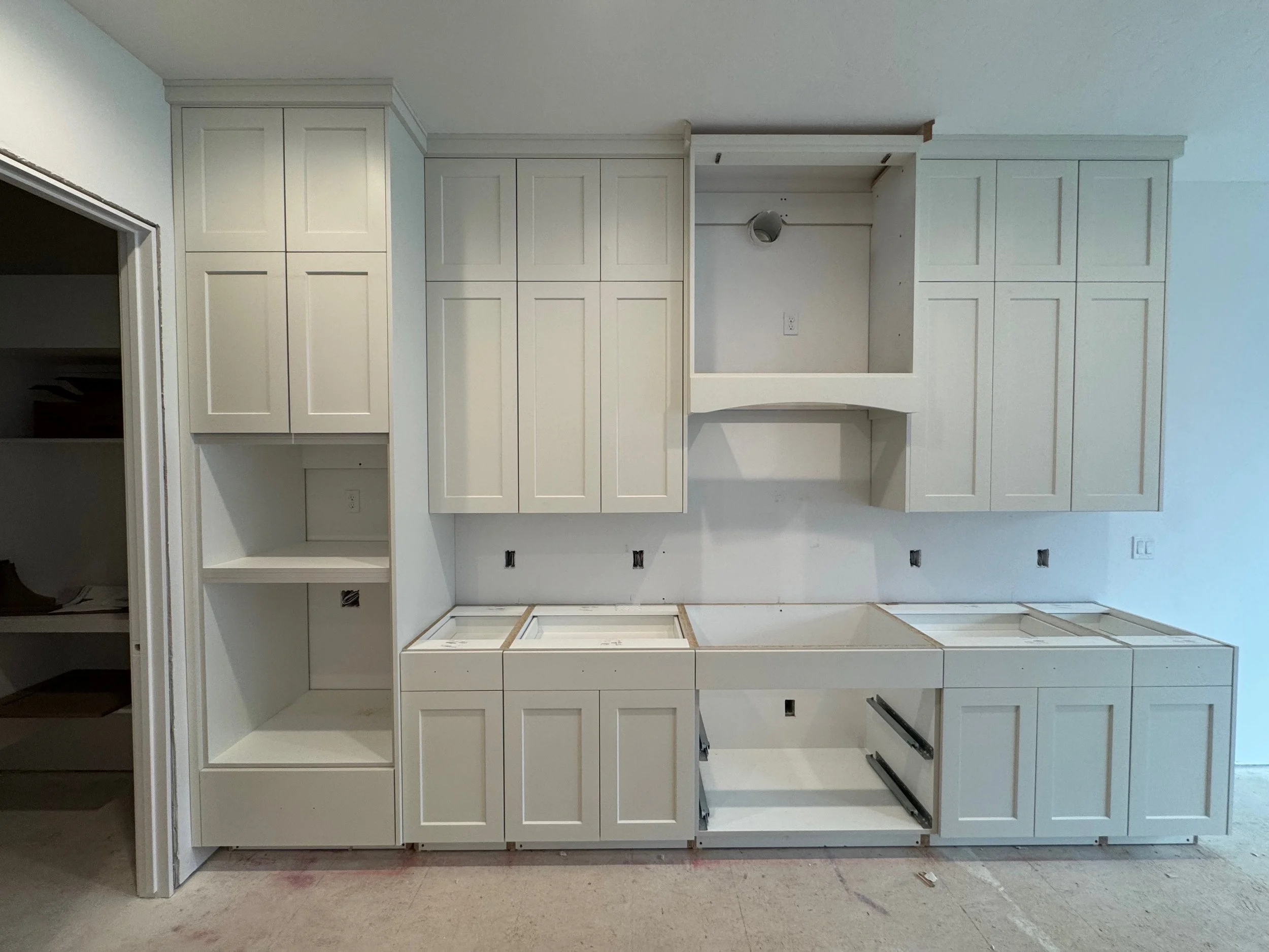 Unfinished kitchen cabinetry with white cabinets, some doors attached and others missing, and an open space for appliances or a sink, with electrical outlets and a concrete floor.