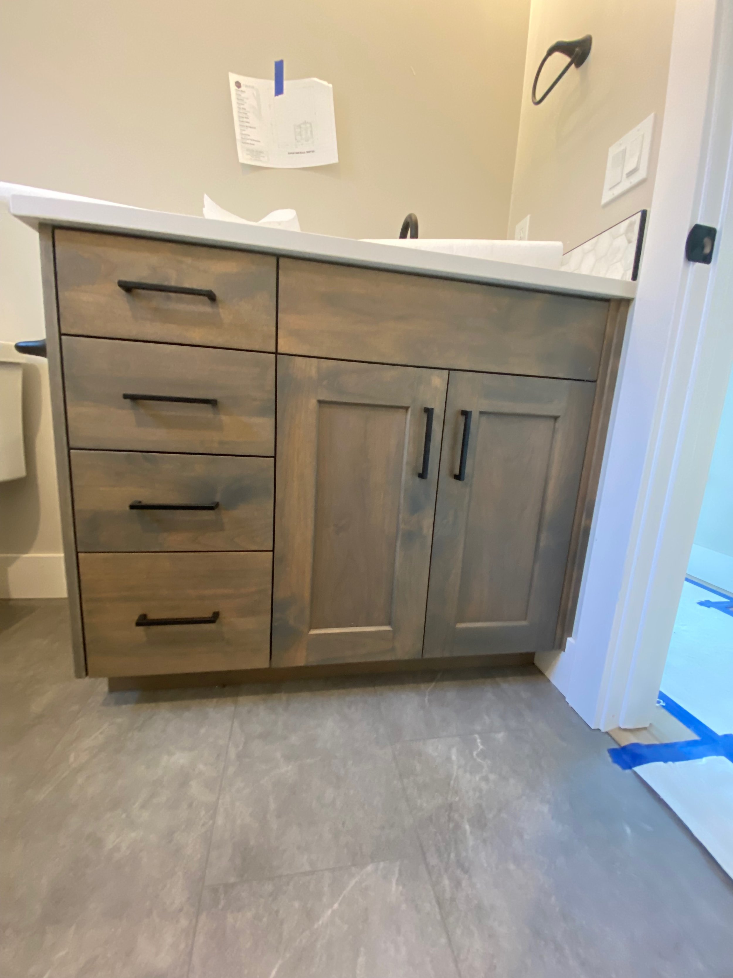 A wooden bathroom vanity with a white countertop, six drawers, and two cabinet doors, in a room with gray tile flooring and beige walls.