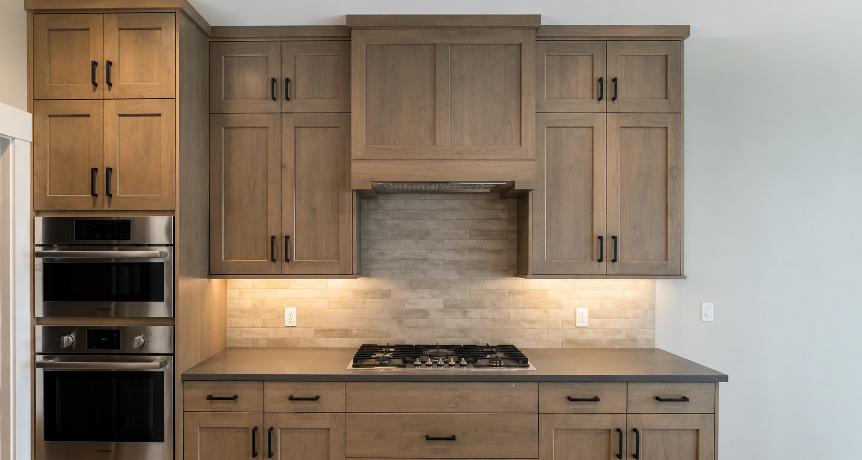 Modern kitchen with wooden cabinets, a built-in double oven, and a stovetop under a range hood. Light-colored subway tile backsplash and under-cabinet lighting.