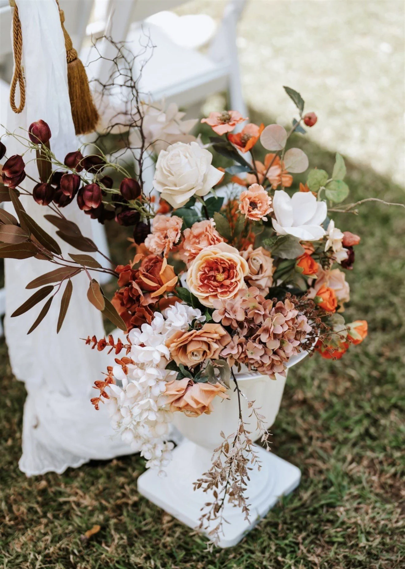A floral arrangement featuring various peach, white, and dark red flowers, with green and reddish leaves, placed on a white pedestal outdoors, with a grassy ground in the background.