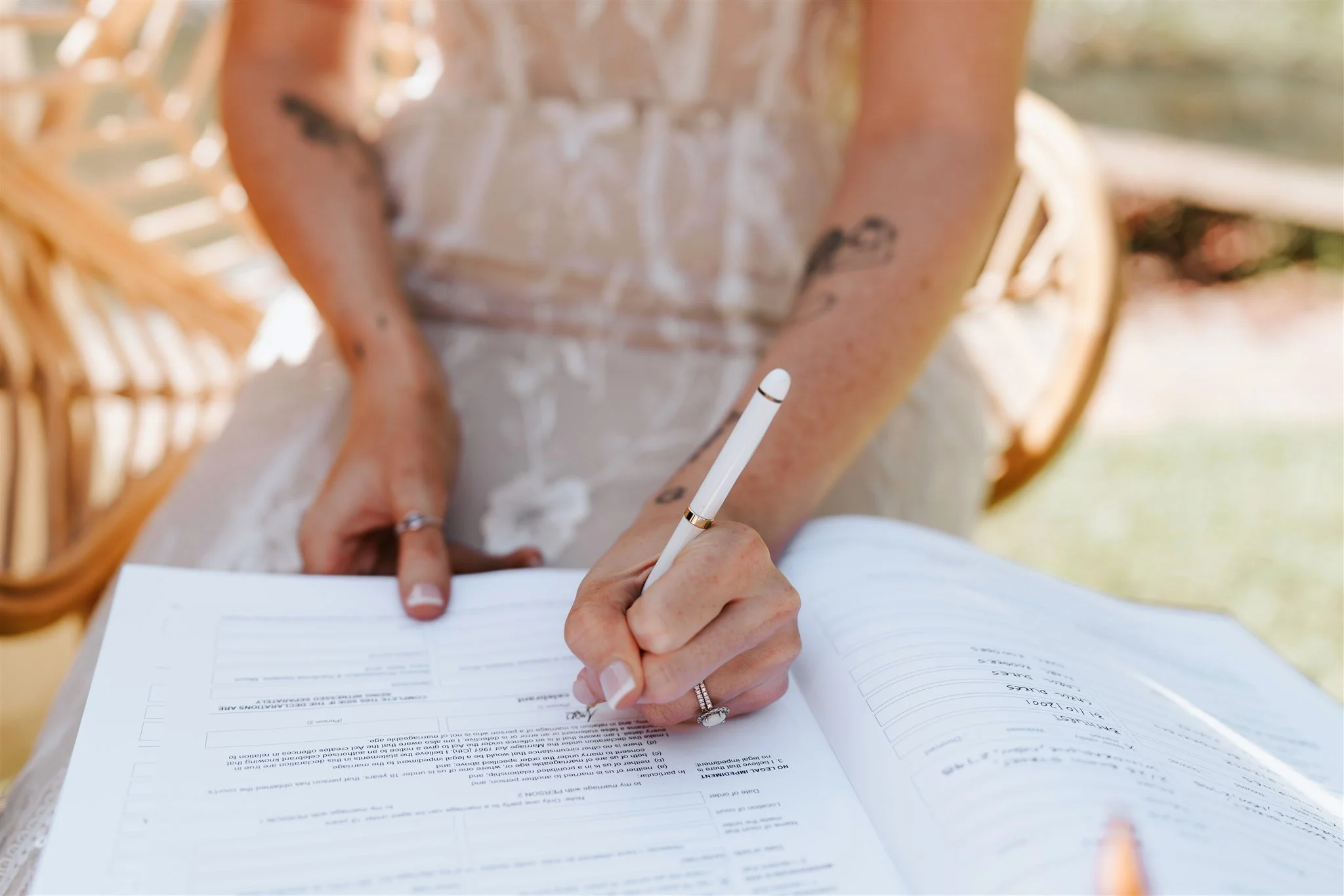 Person signing documents with a lace dress, rings, and a white pen, at a table outside.