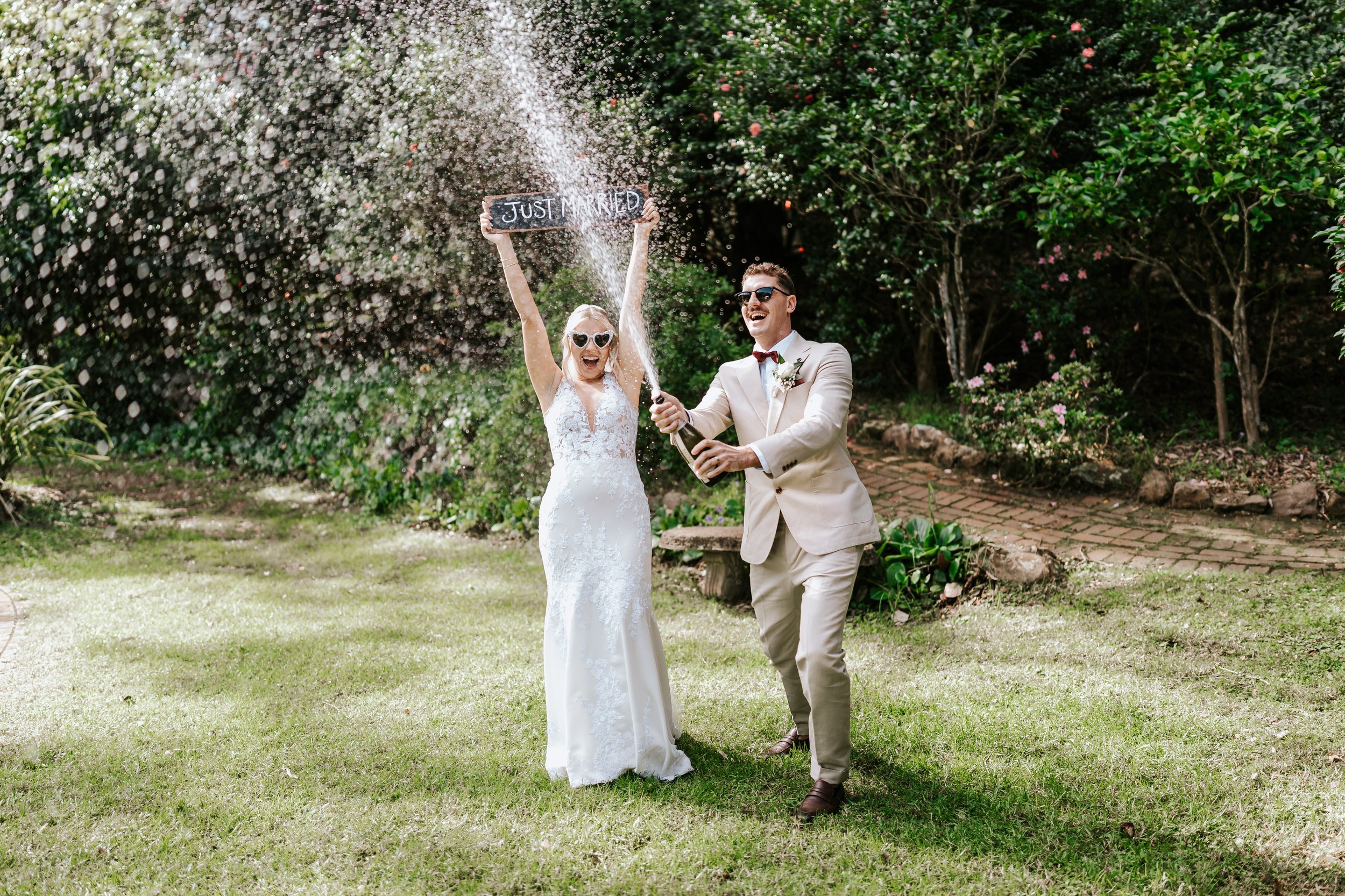 bride and groom joyfully celebrate their marriage with a bottle of champagne and a Just Married sign in a garden setting
