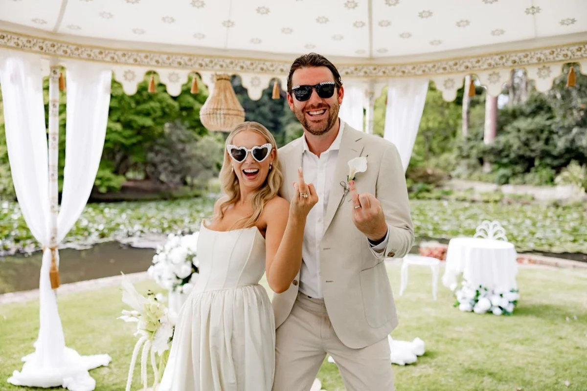 A happy couple at their wedding, both wearing sunglasses and forming middle finger gestures, standing under a white gazebo with green outdoor scenery in the background.