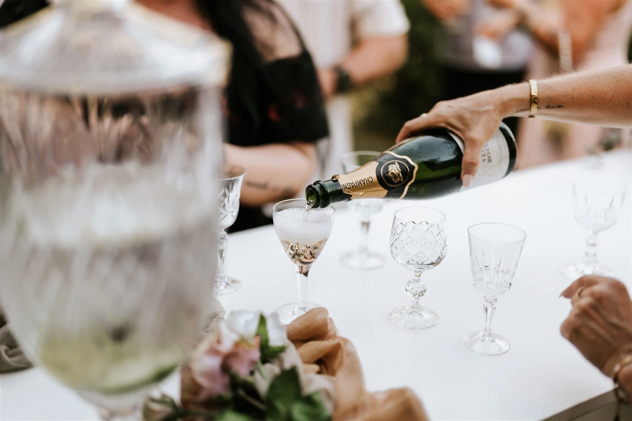 A person pouring champagne into a glass at a celebration or gathering, with blurred people and floral arrangements in the background.
