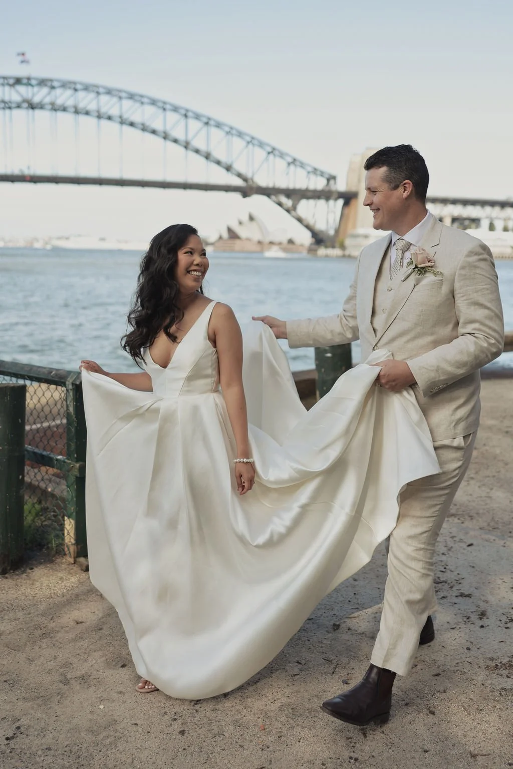 A bride and groom on a beach near the Sydney Harbour Bridge, smiling at each other. The bride is in a satin wedding dress, and the groom is in a light-colored suit, holding the bride's dress.