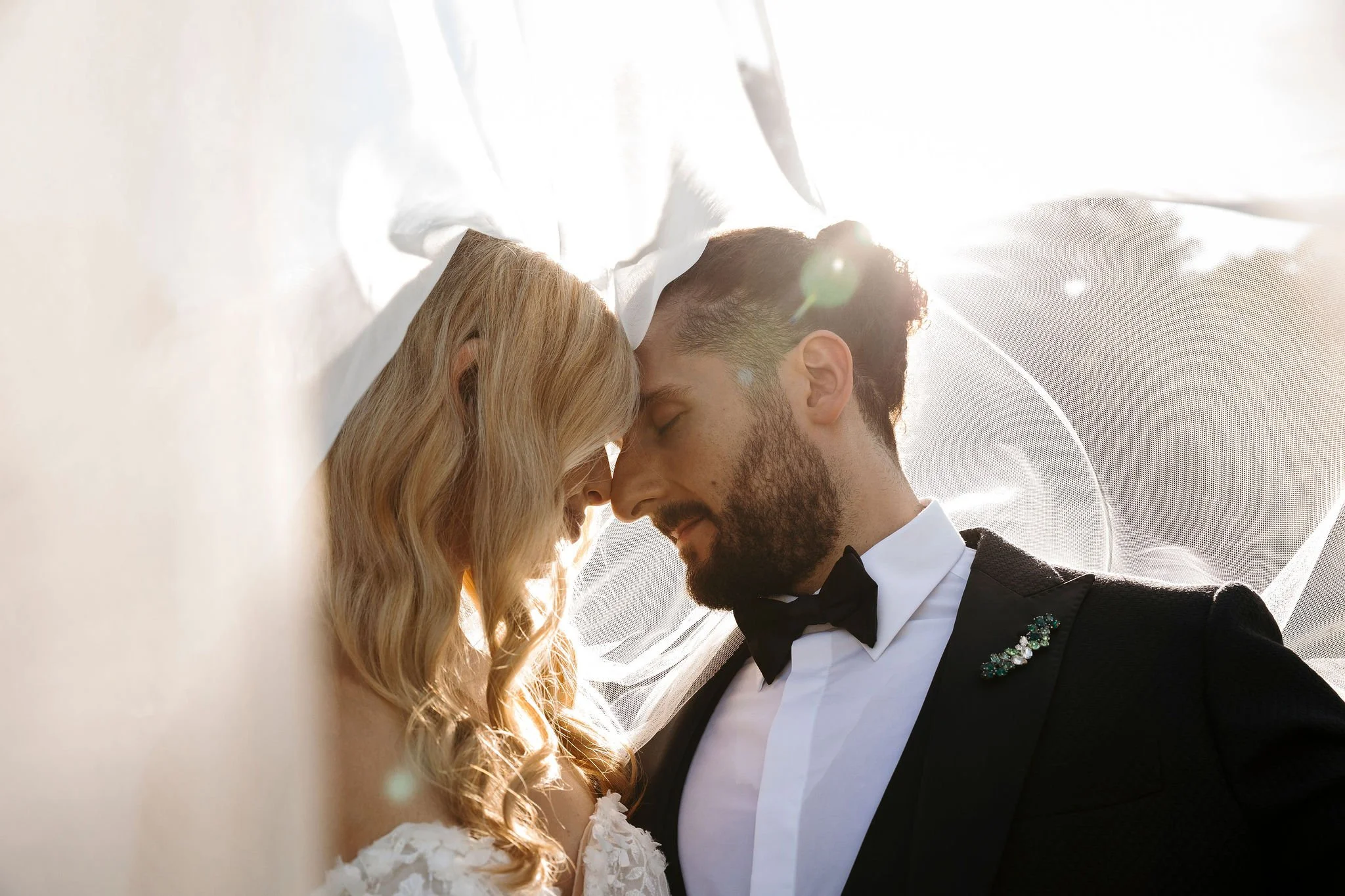 A bride and groom are closely leaning foreheads together, eyes closed, on their wedding day. The bride has long, wavy blonde hair and is wearing a white dress with lace details, and the groom has short hair and a beard, wearing a black tuxedo with a white shirt and black bow tie. Bright sunlight creates a backlit effect around them.