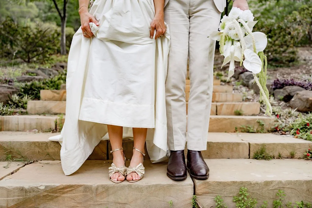 Close-up of a bride and groom standing on stone steps, holding white flowers, with the focus on their lower bodies and attire.