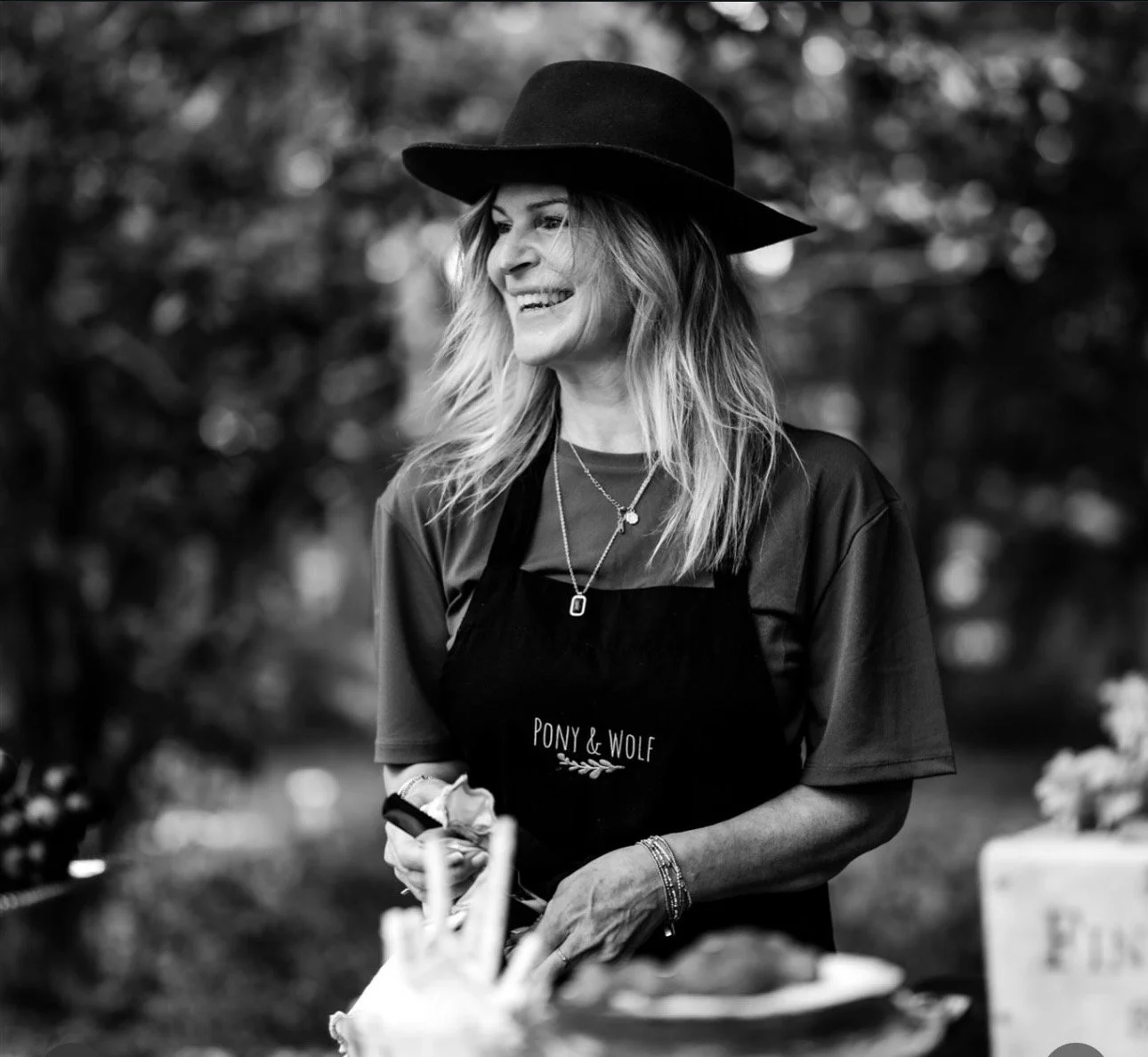A woman wearing a wide-brimmed hat and an apron with the words "Pony & Wolf" smiles outdoors, with blurred trees in the background.
