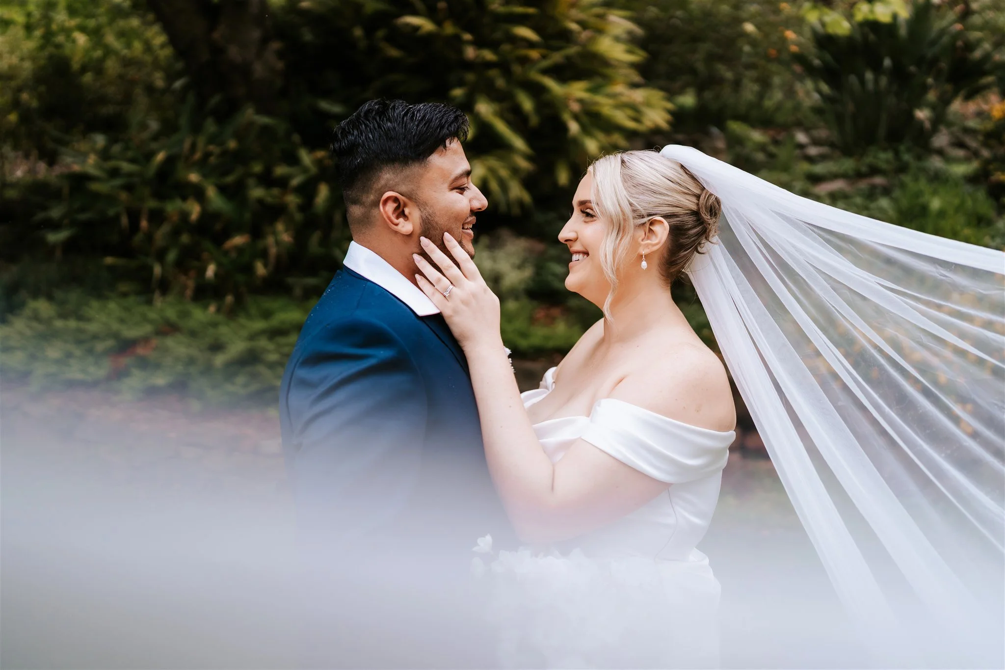 A bride and groom smiling at each other outdoors, with the bride touching the groom's face and their foreheads nearly touching. The bride is wearing earrings and a veil, and the groom is dressed in a dark suit. There is greenery in the background.