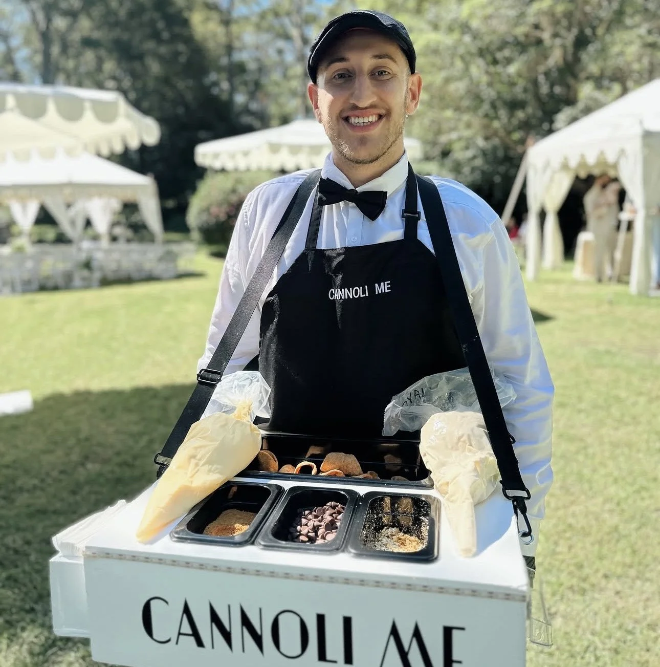 A smiling man dressed as a waiter, wearing a tuxedo with a bow tie and apron that says 'CANNOLI ME,' serving cannoli and toppings from a mobile cart outdoors at a garden event with white tents in the background.