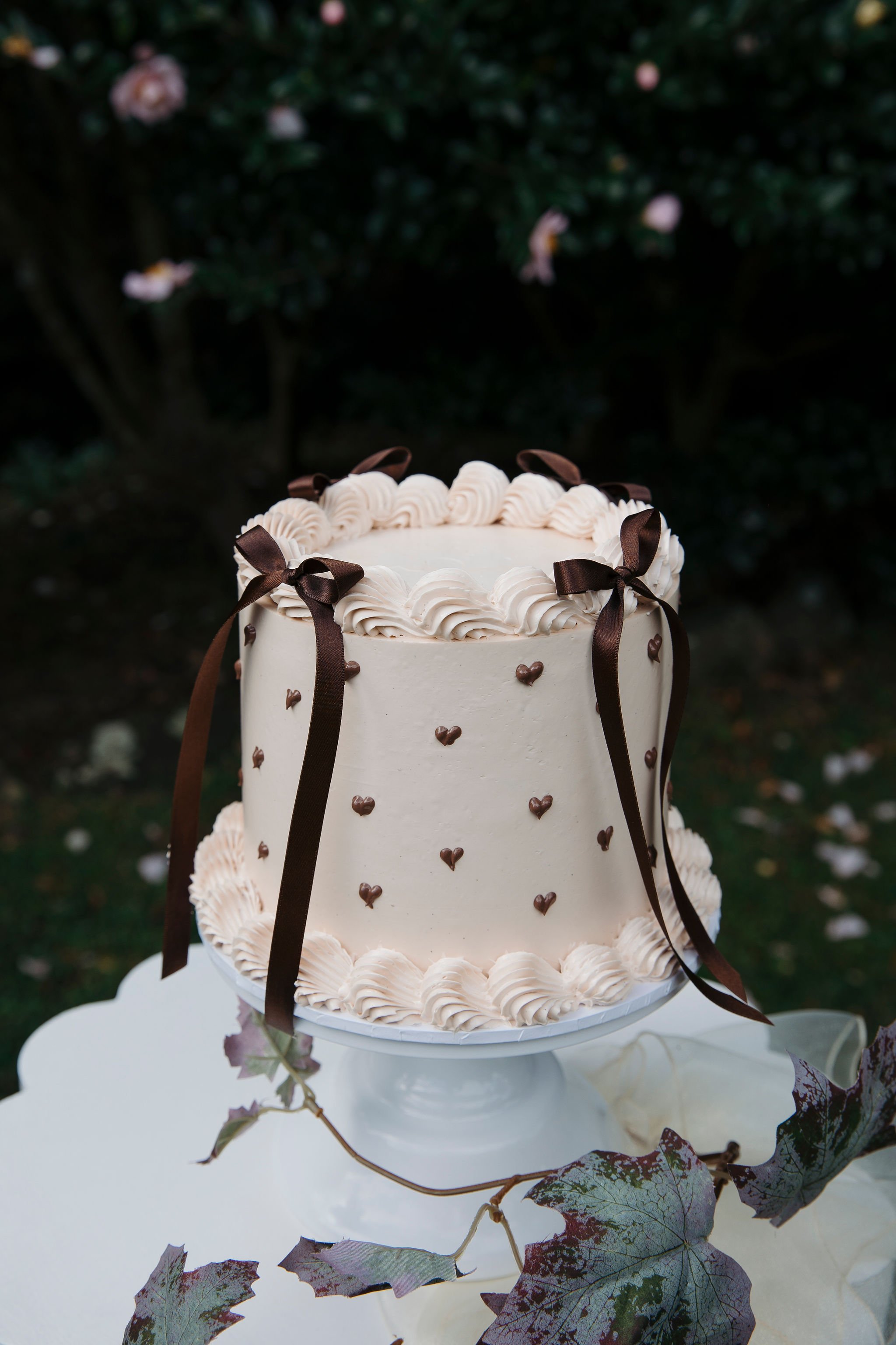 A tall, tiered wedding cake decorated with small chocolate hearts and brown ribbons, topped with swirls of white cream, standing outdoors in front of a dark green leafy background, surrounded by purple and green leaves.