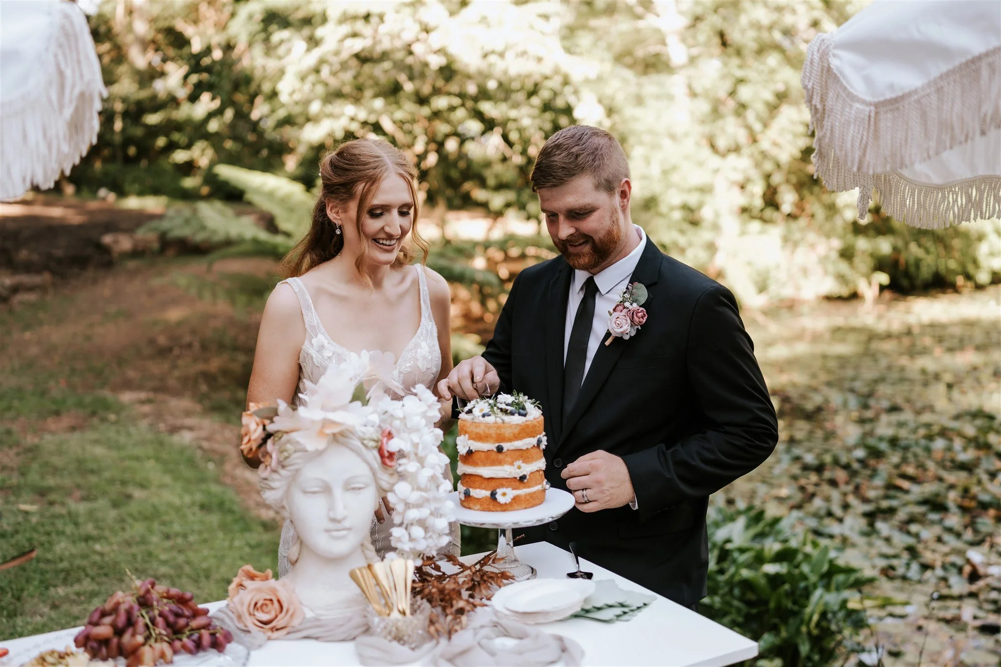 A bride and groom celebrating at an outdoor wedding reception, cutting a layered cake decorated with flowers, with a table of desserts and decorative sculptures in the foreground.