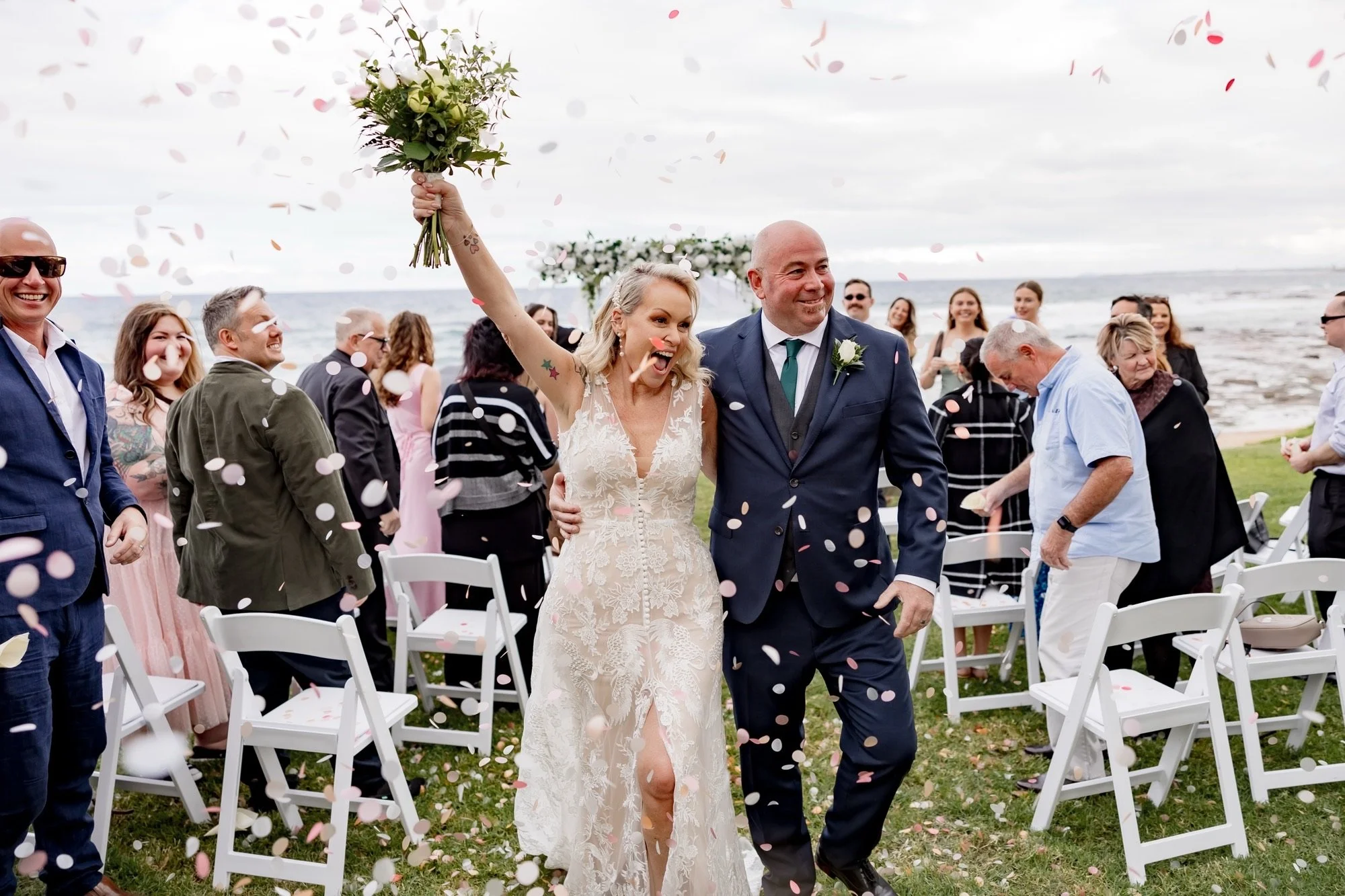 A bride and groom walking down the aisle at an outdoor beach wedding, with guests celebrating around them, pink and white flower petals falling from above, and the ocean in the background.