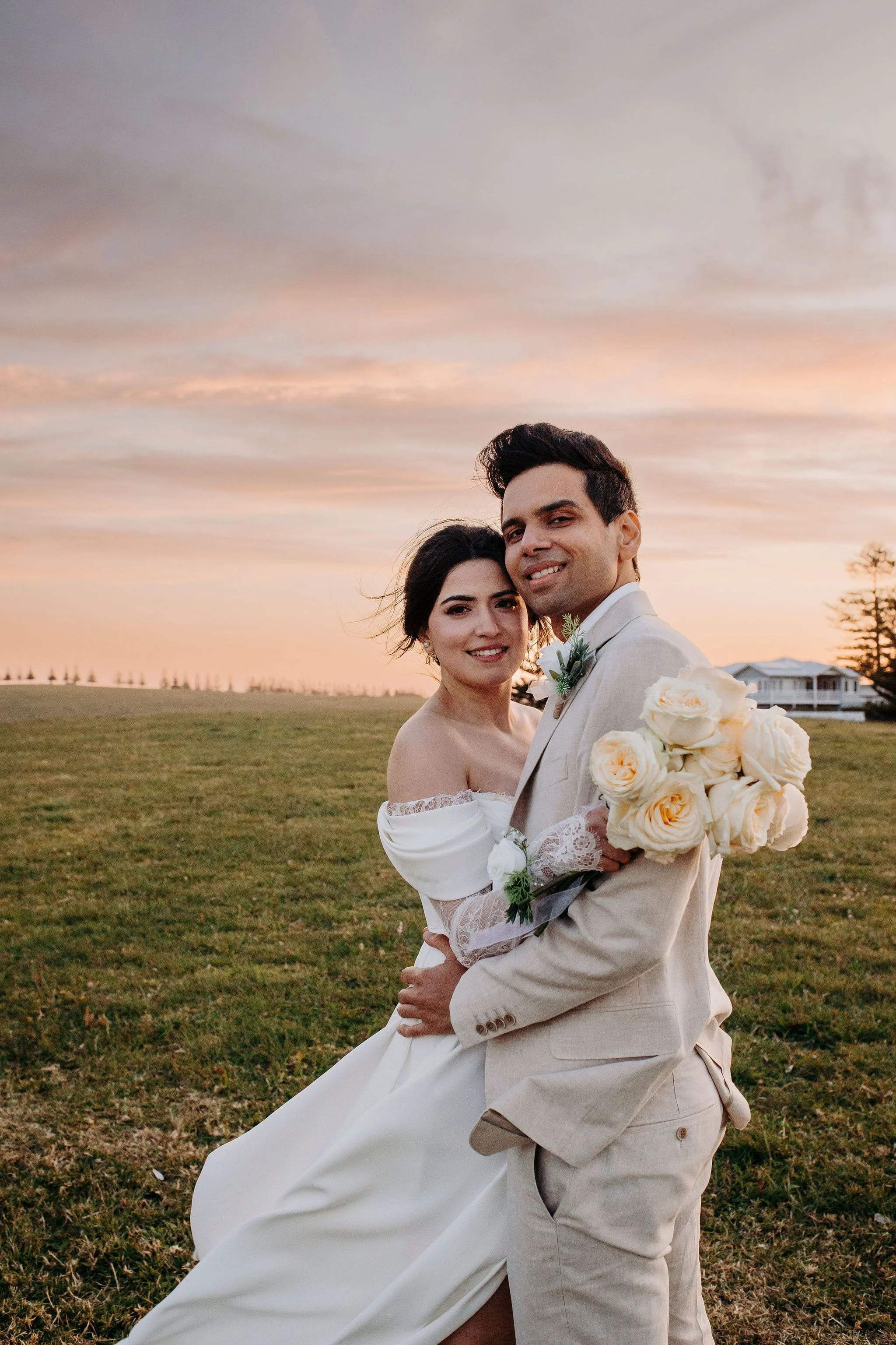 A bride and groom are embracing outdoors at sunset. The bride is holding a bouquet of cream roses, and they are both smiling. The background features an open field with a house in the distance and a colorful sky.