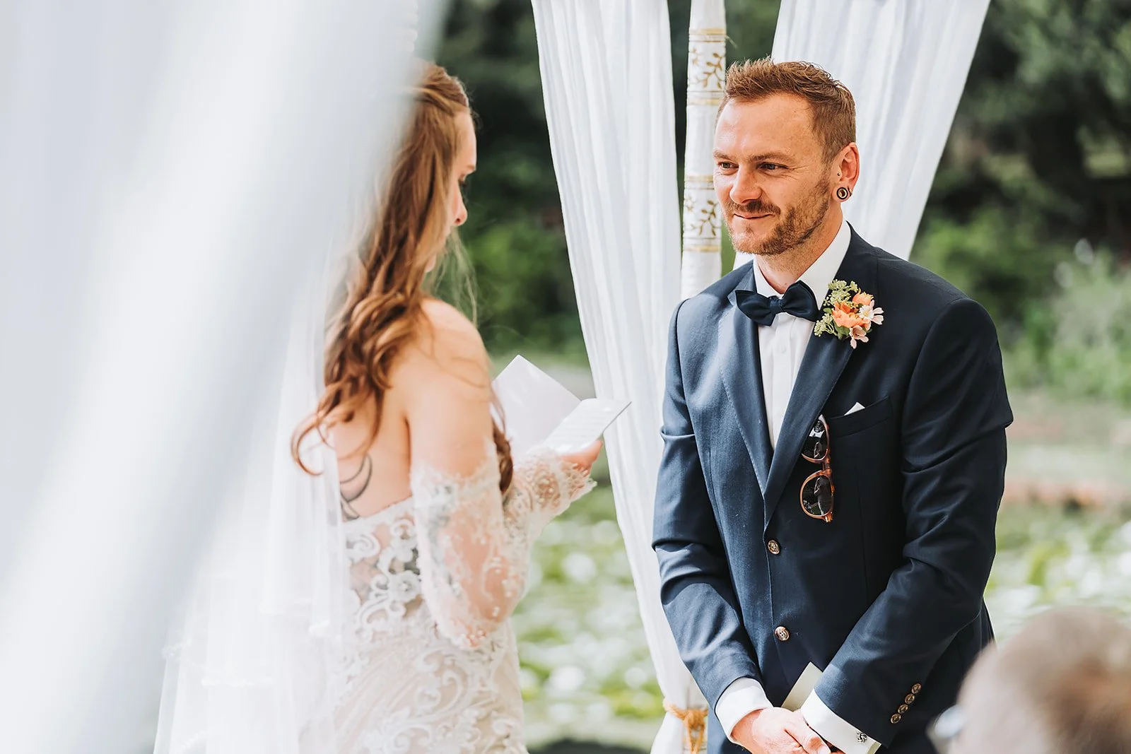 A groom with a bow tie and a boutonniere faces a bride with long, wavy hair reading from a paper during a wedding ceremony outdoors, framed by white curtains.