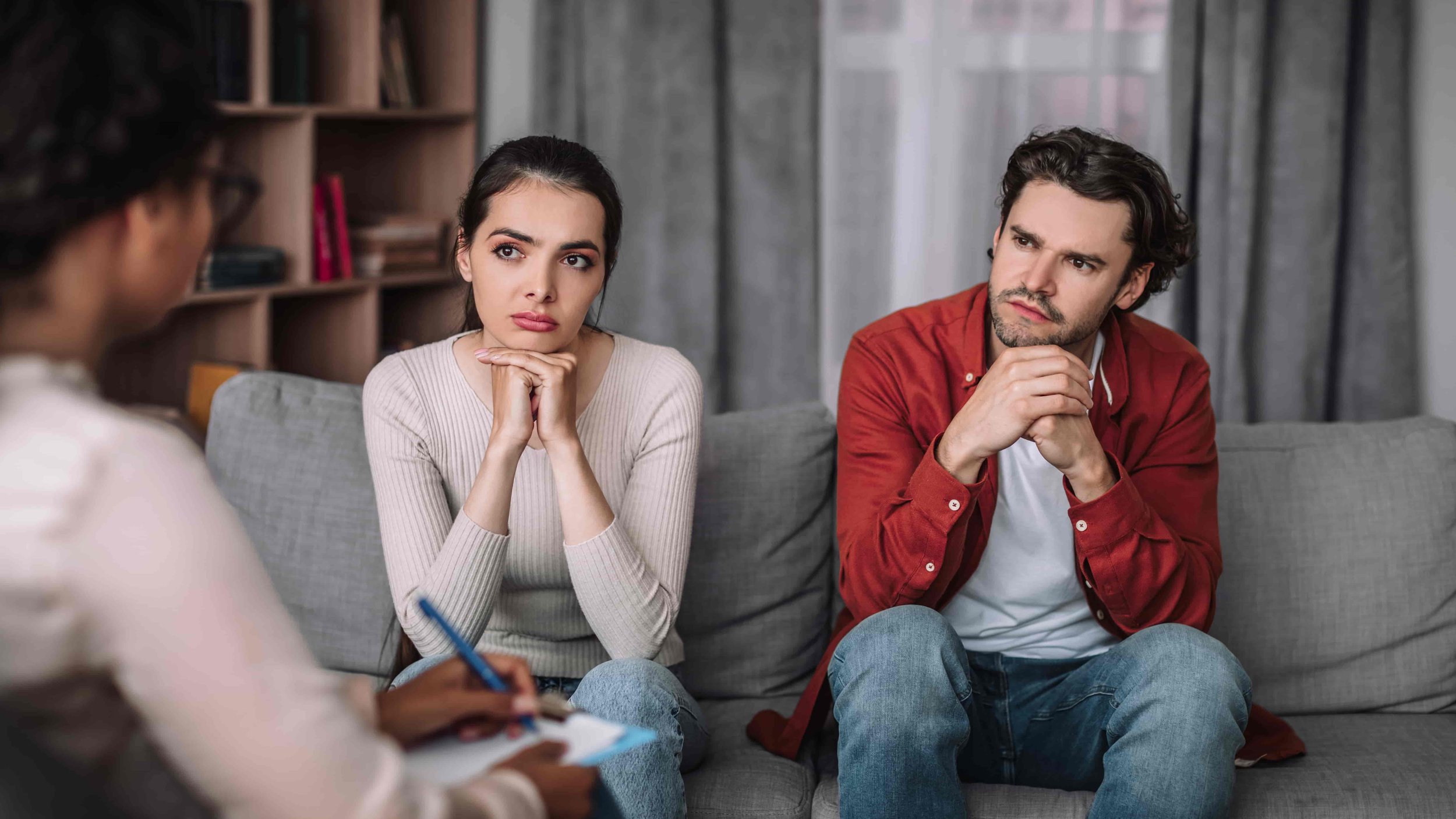 A woman with dark hair and a man with brown curly hair sitting on a gray couch, listening attentively to someone in a counseling room with a bookshelf and gray curtains.