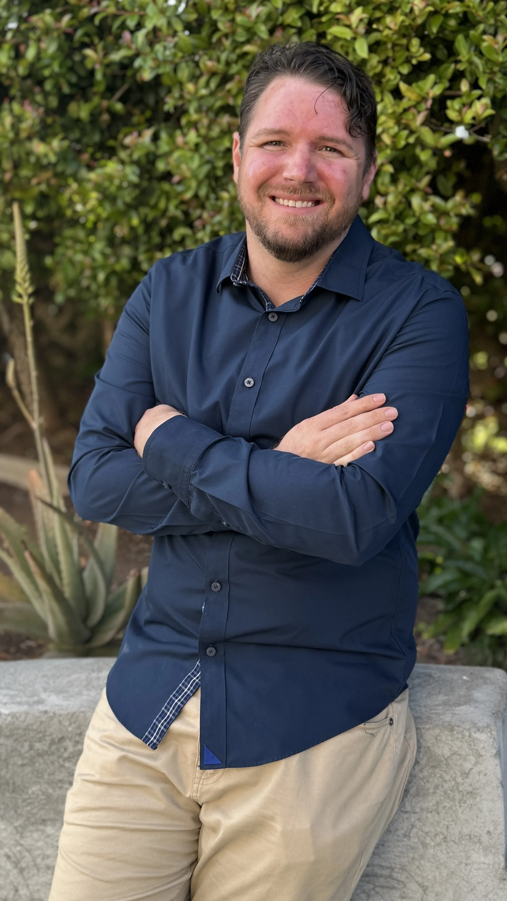 Justin Latimer, a man with light skin, dark hair, and a beard, smiling with arms crossed, wearing a navy blue button-up shirt and beige pants, standing outdoors in front of green bushes.