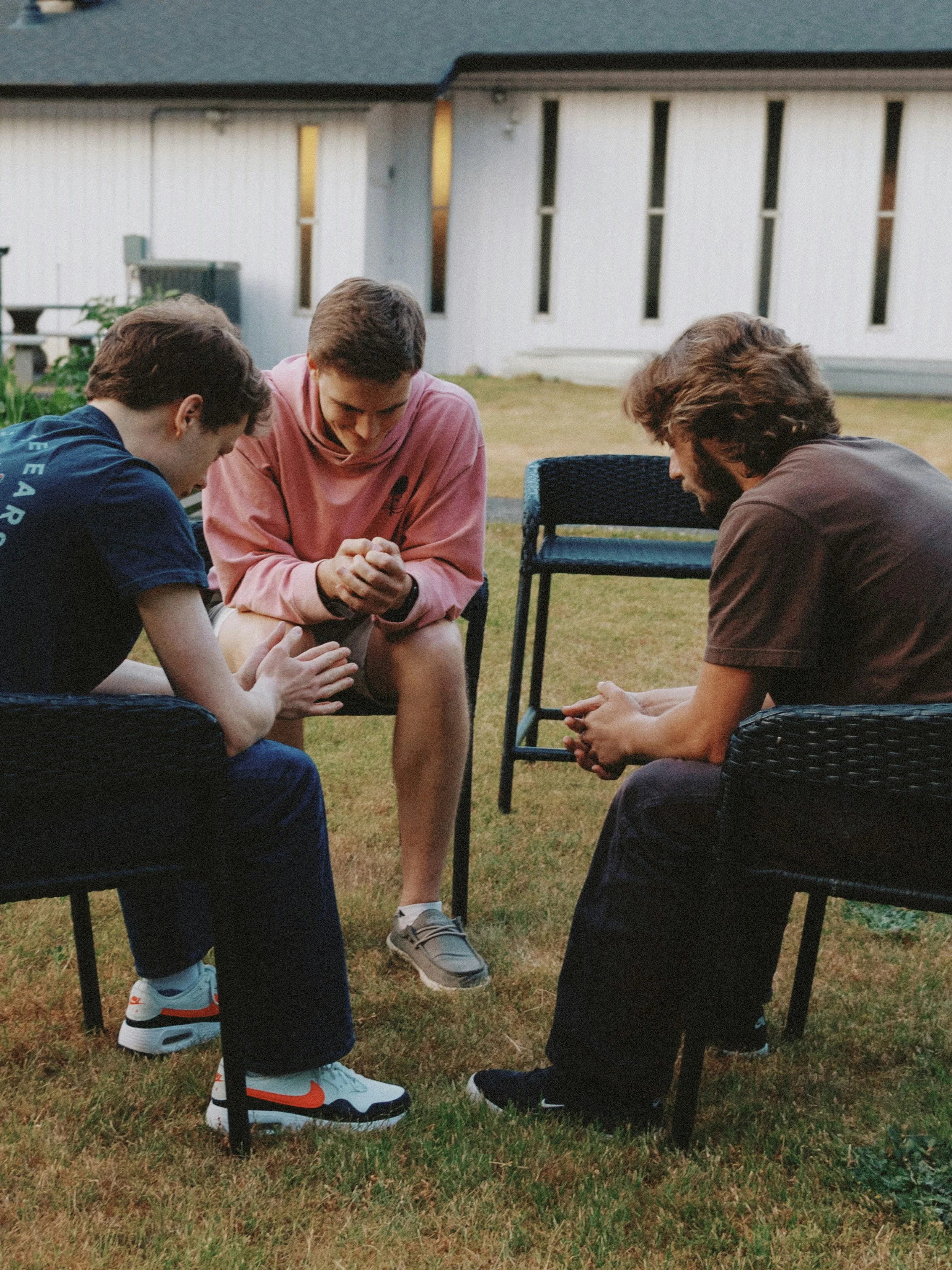 Three young men are kneeling and bowing their heads with clasped hands in a circle outdoors on grass, with a white building and picket fence in the background during dusk.