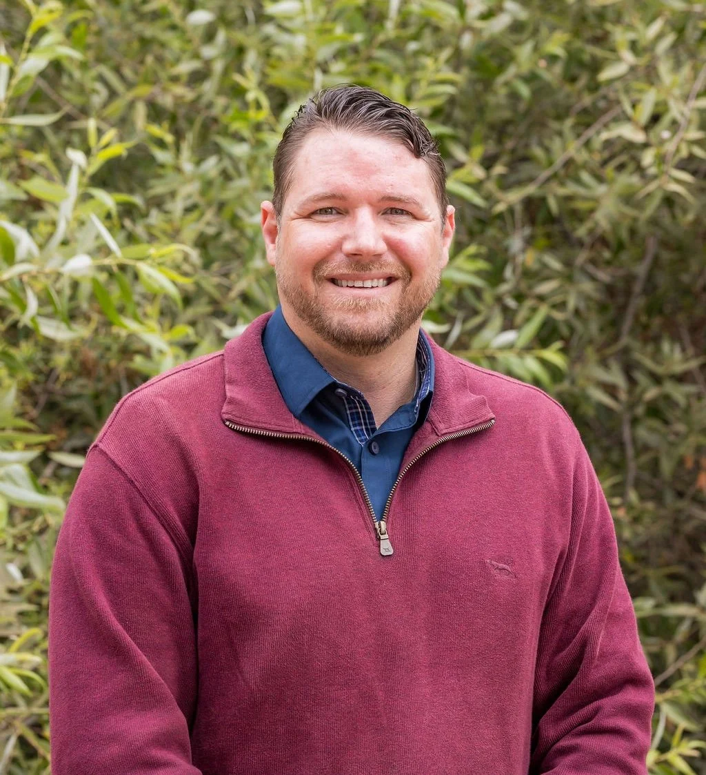 Justin Latimer, a man smiling outdoors in front of green bushes, wearing a burgundy quarter-zip sweater over a blue collared shirt.