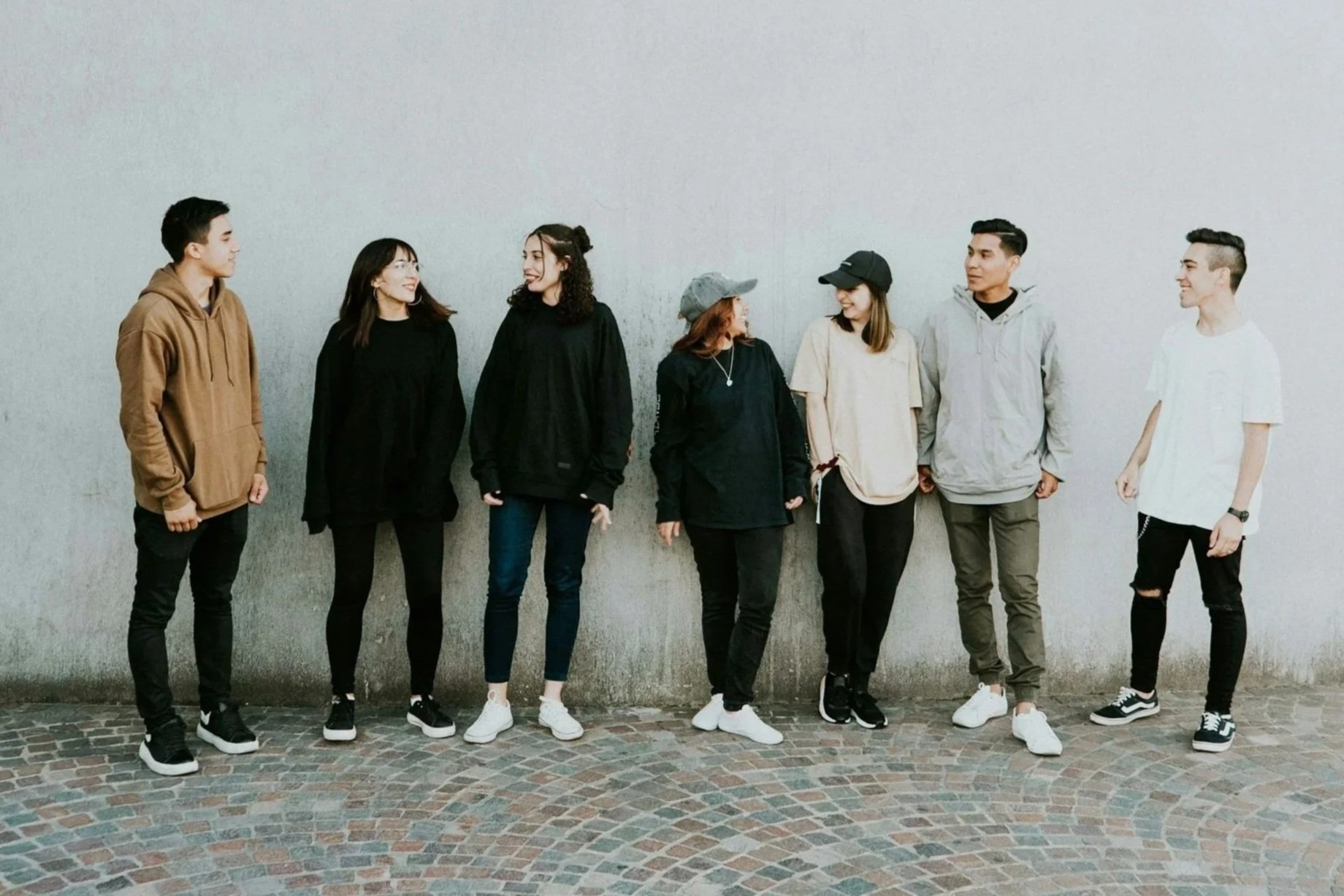 Seven young people standing against a plain gray wall, engaged in conversation and smiling, dressed casually with sneakers and streetwear.