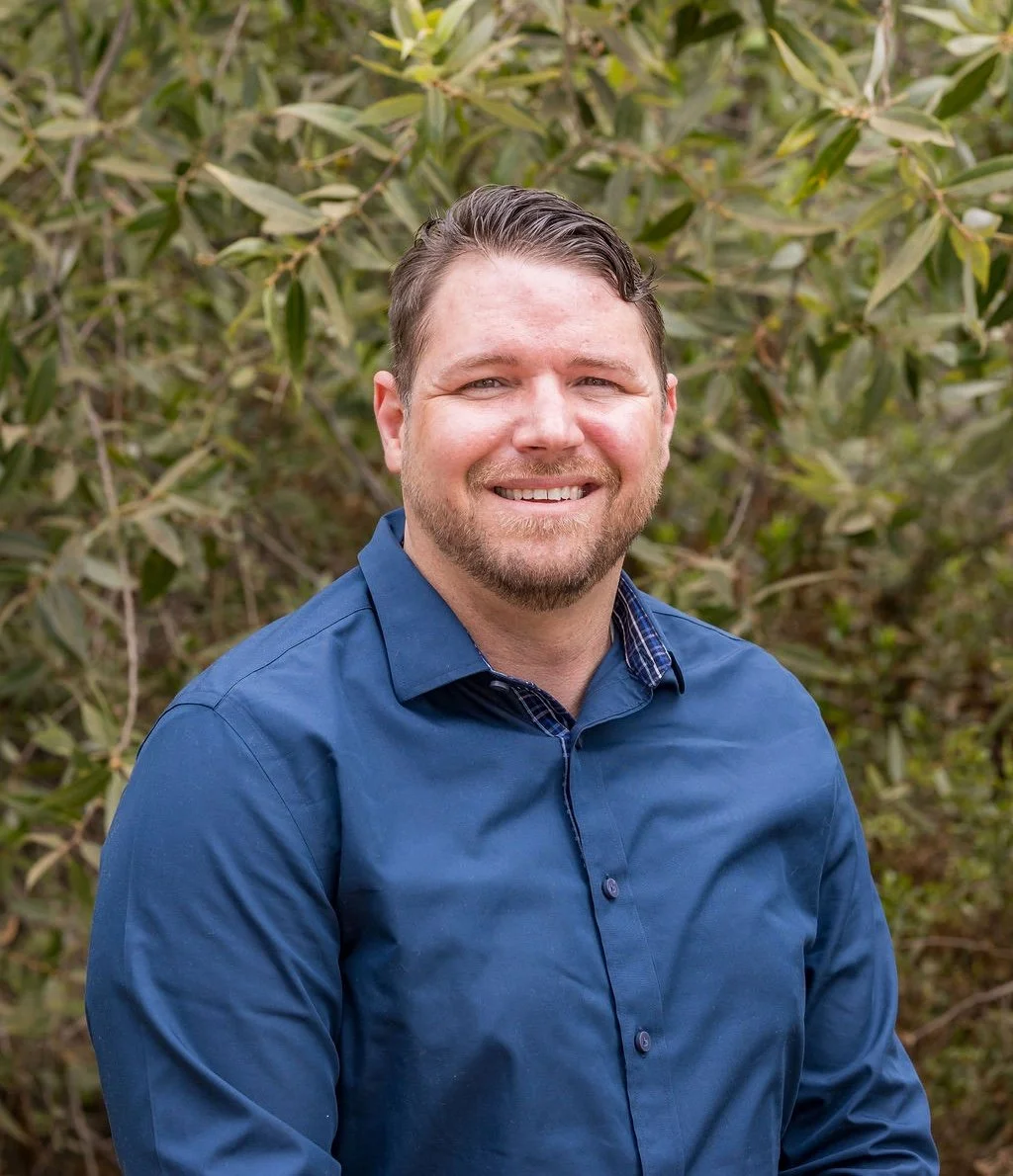 Justin Latimer, a man with short dark hair, beard, and mustache, smiling in front of green foliage, wearing a blue button-up shirt.