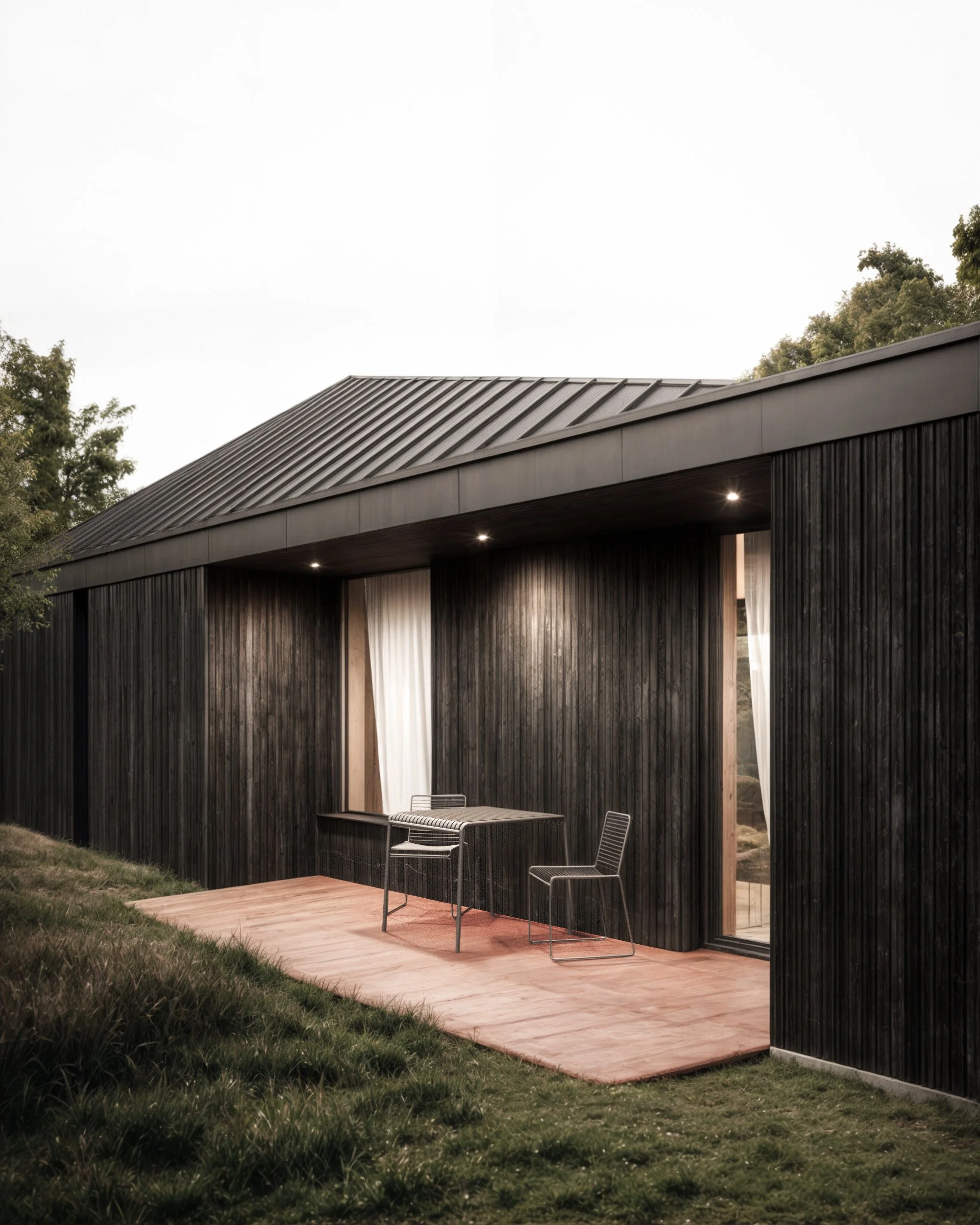 Modern black wooden house with a metal roof, outdoor patio with a small table and chairs, surrounded by green grass and trees, during evening with subtle lighting.