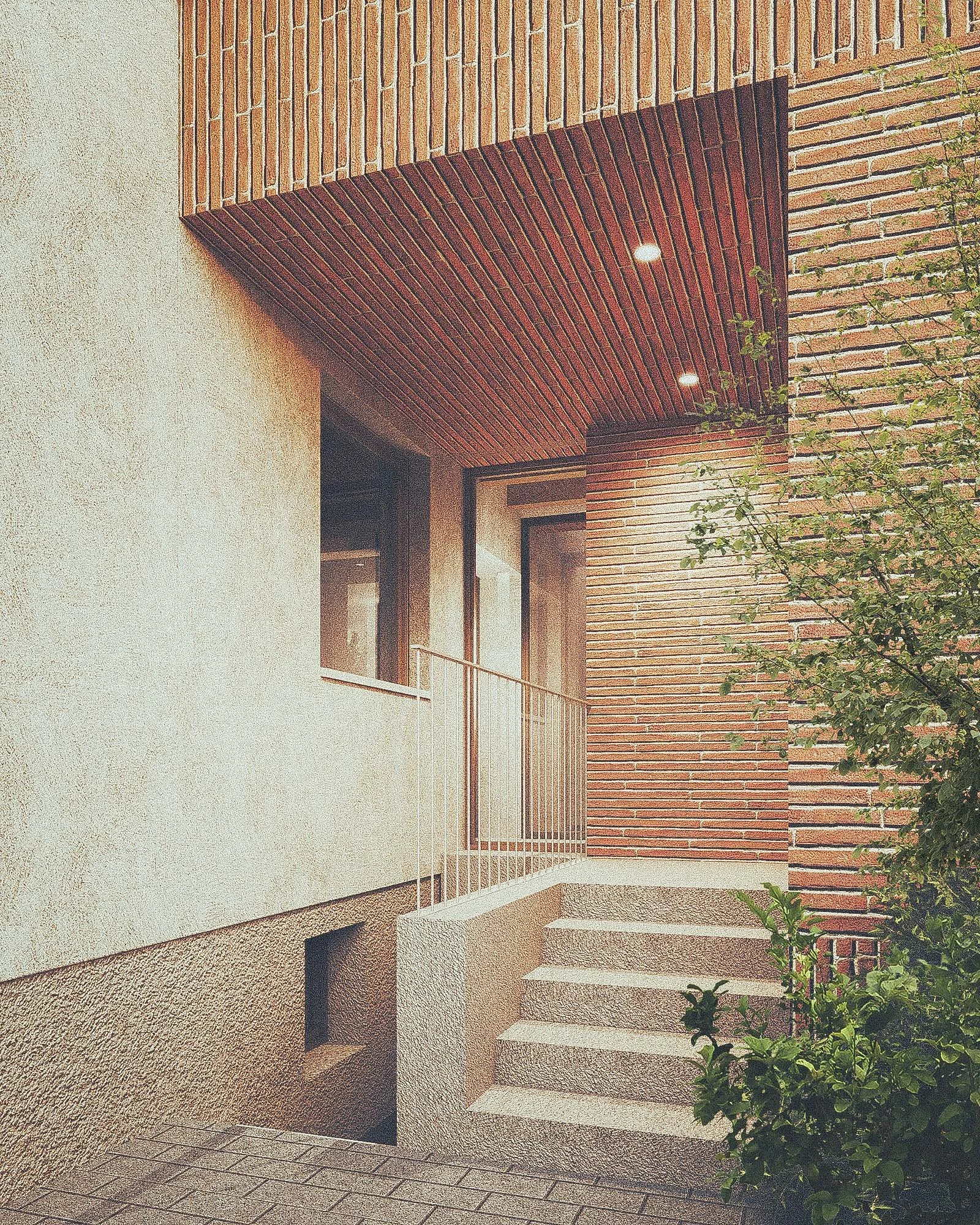 Exterior view of a modern building with a brick facade and a balcony. The structure has a mix of smooth white and brick walls, with lighting in the ceiling of the balcony area. There is a small garden plant on the right side of the image.
