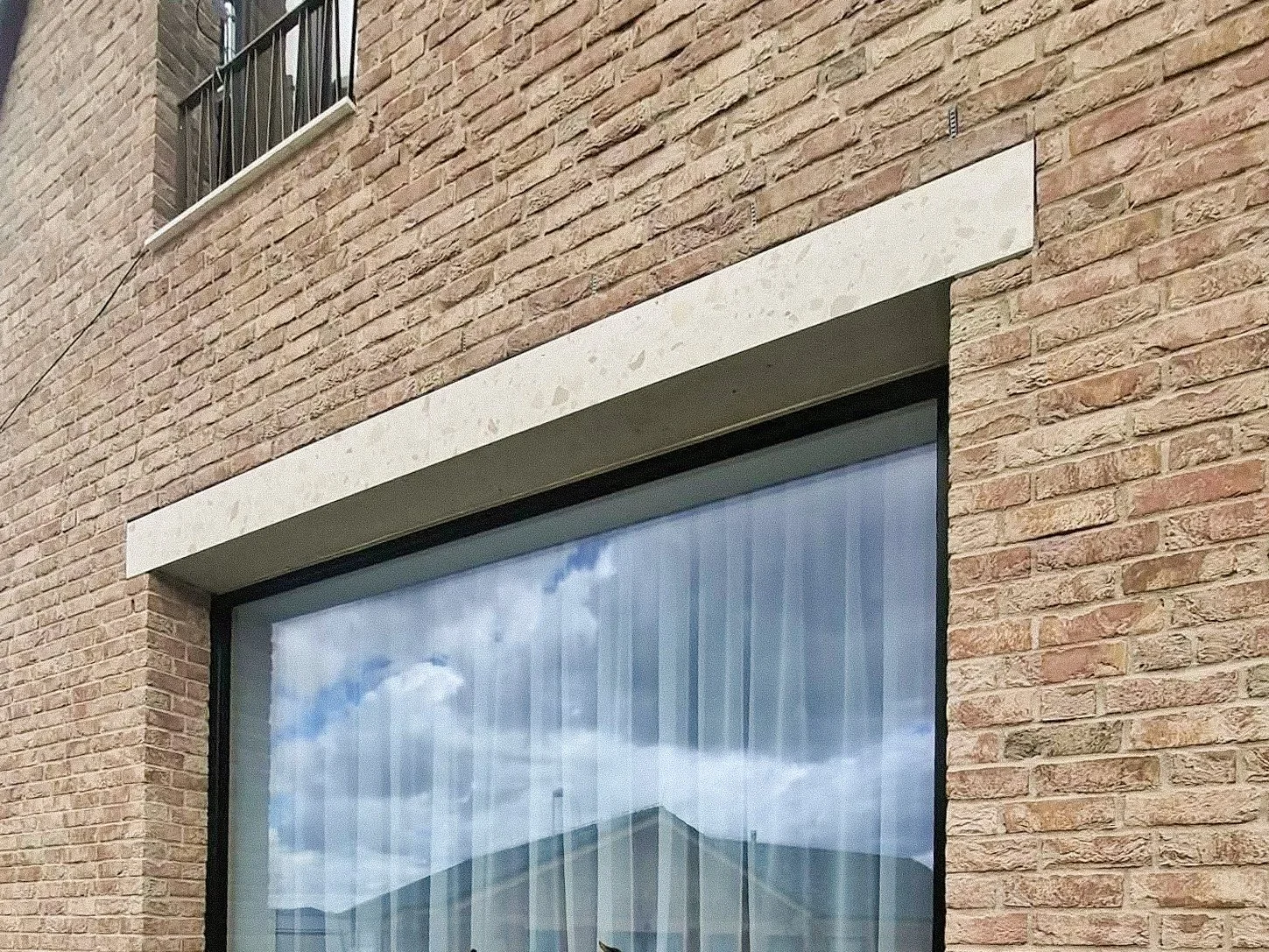 Close-up of a brick building wall with a large window reflecting the cloudy sky and part of the roofline.