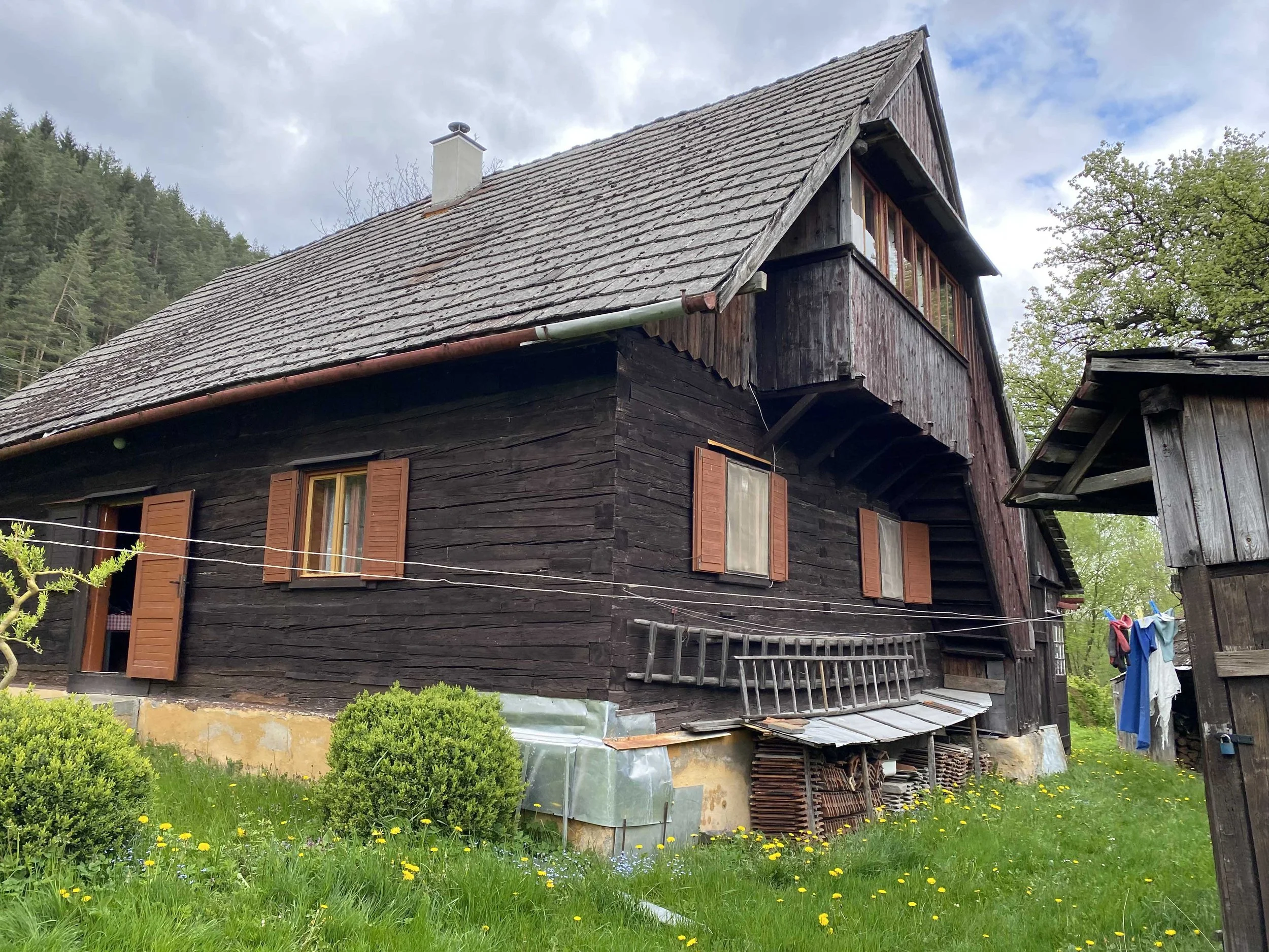 A rustic, wooden house with a steep, tiled roof and open windows, surrounded by greenery and laundry hanging outside.