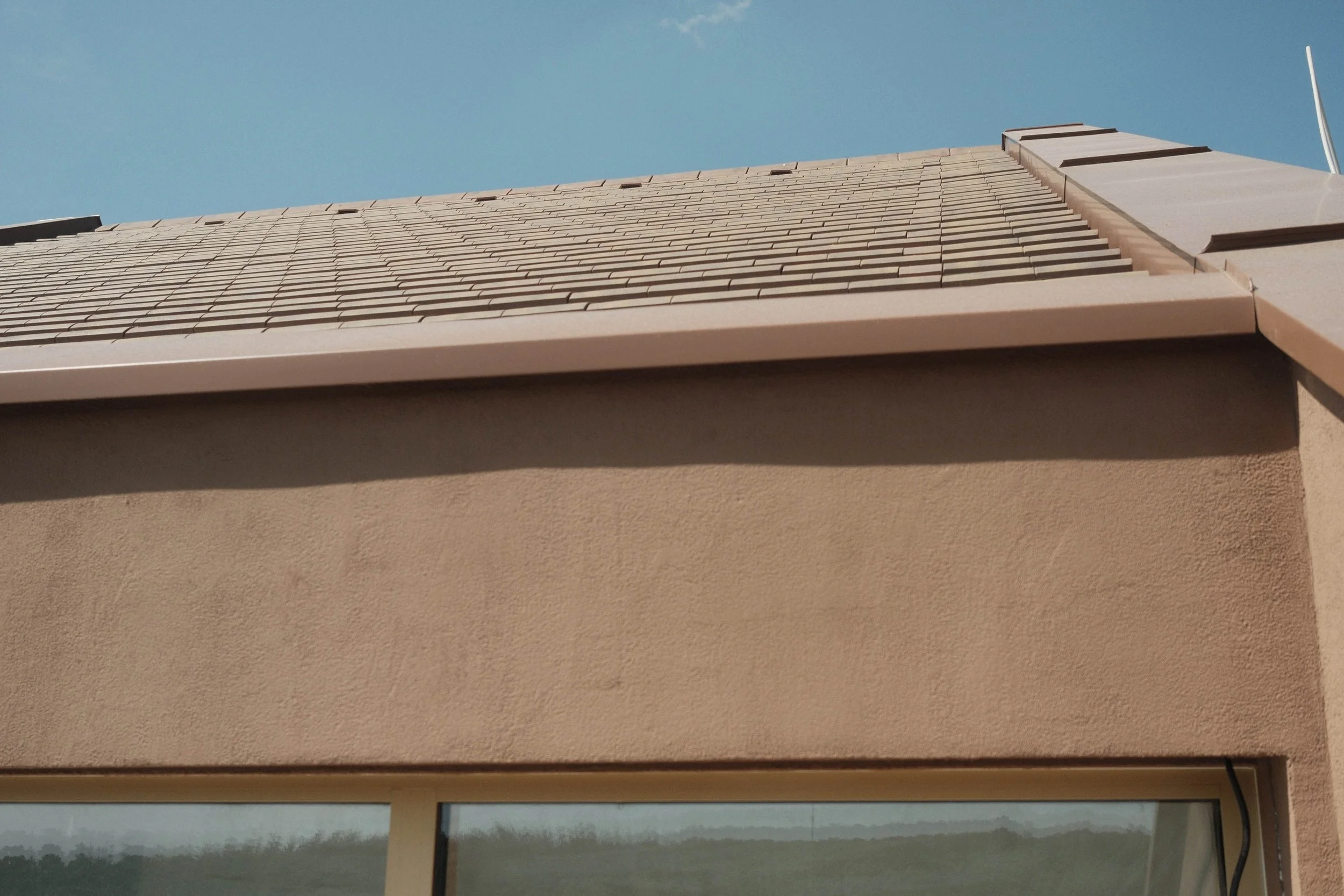 Close-up view of a house roof with beige shingles and a small portion of a window below, against a clear blue sky.