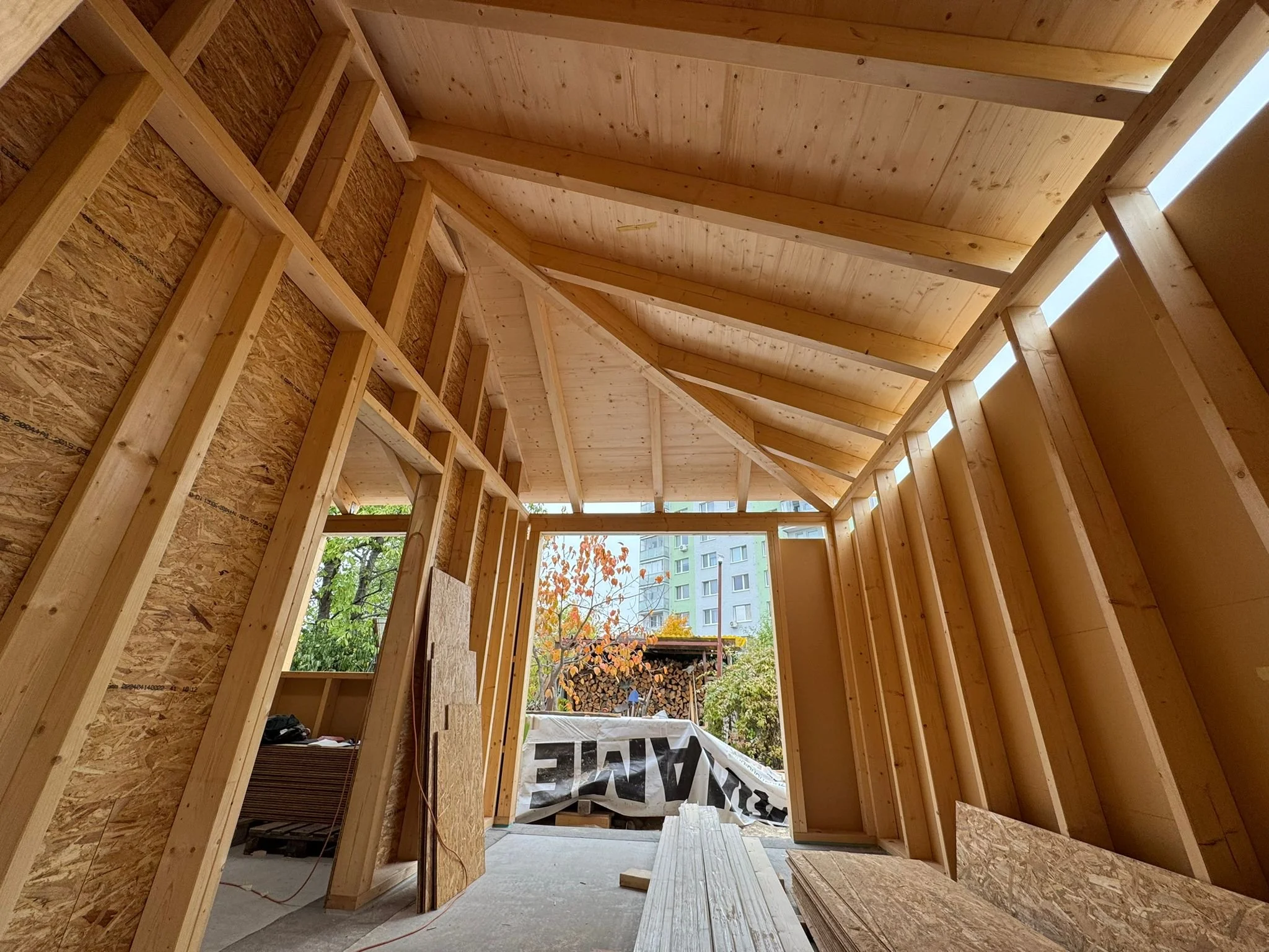 Inner wooden structure of a building under construction, with exposed wooden beams, walls, and ceiling, open to the outdoors showing trees and nearby buildings.
