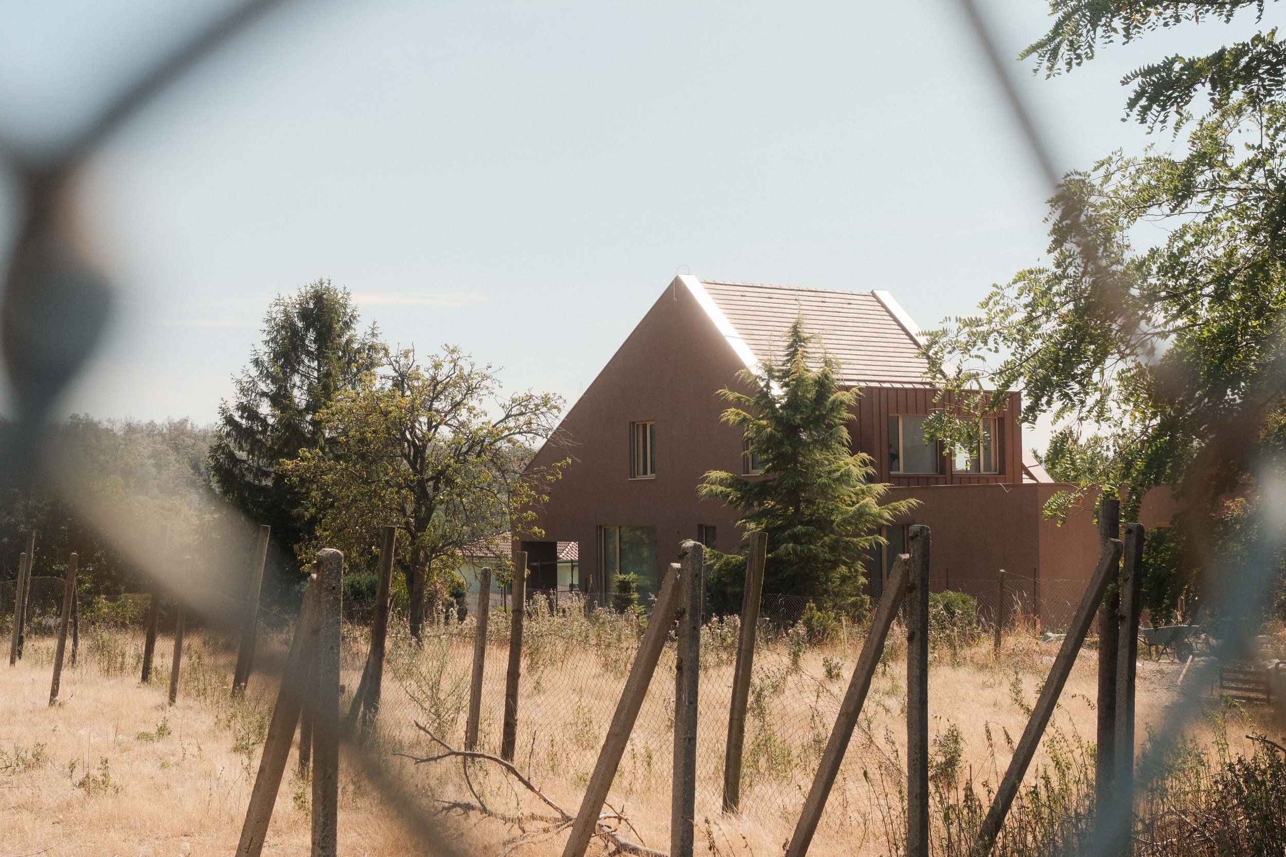 A modern house with a triangular roof, surrounded by trees and enclosed by a fence, viewed through a blurred wire fence.