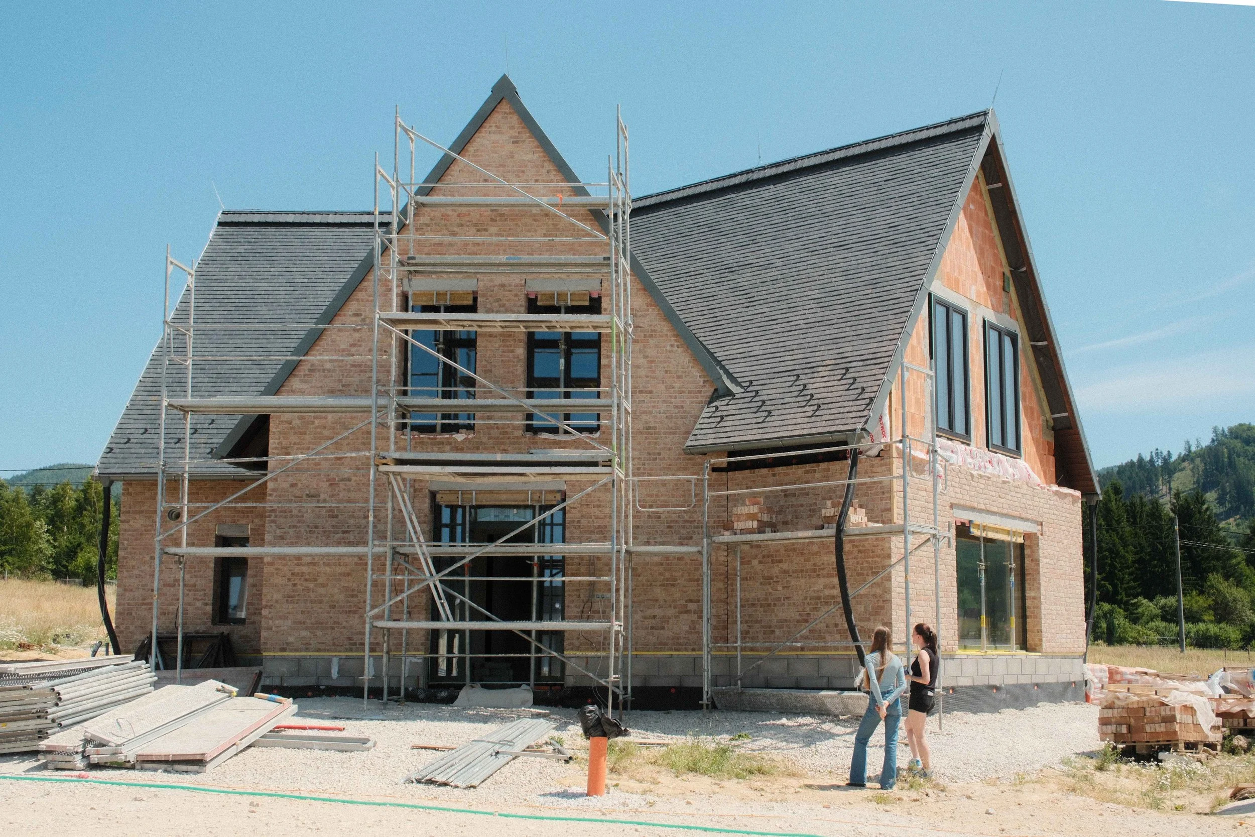 A house under construction with scaffolding, brick walls, and a pitched roof, located in a rural area with trees and hills in the background, and two people standing near the construction site.