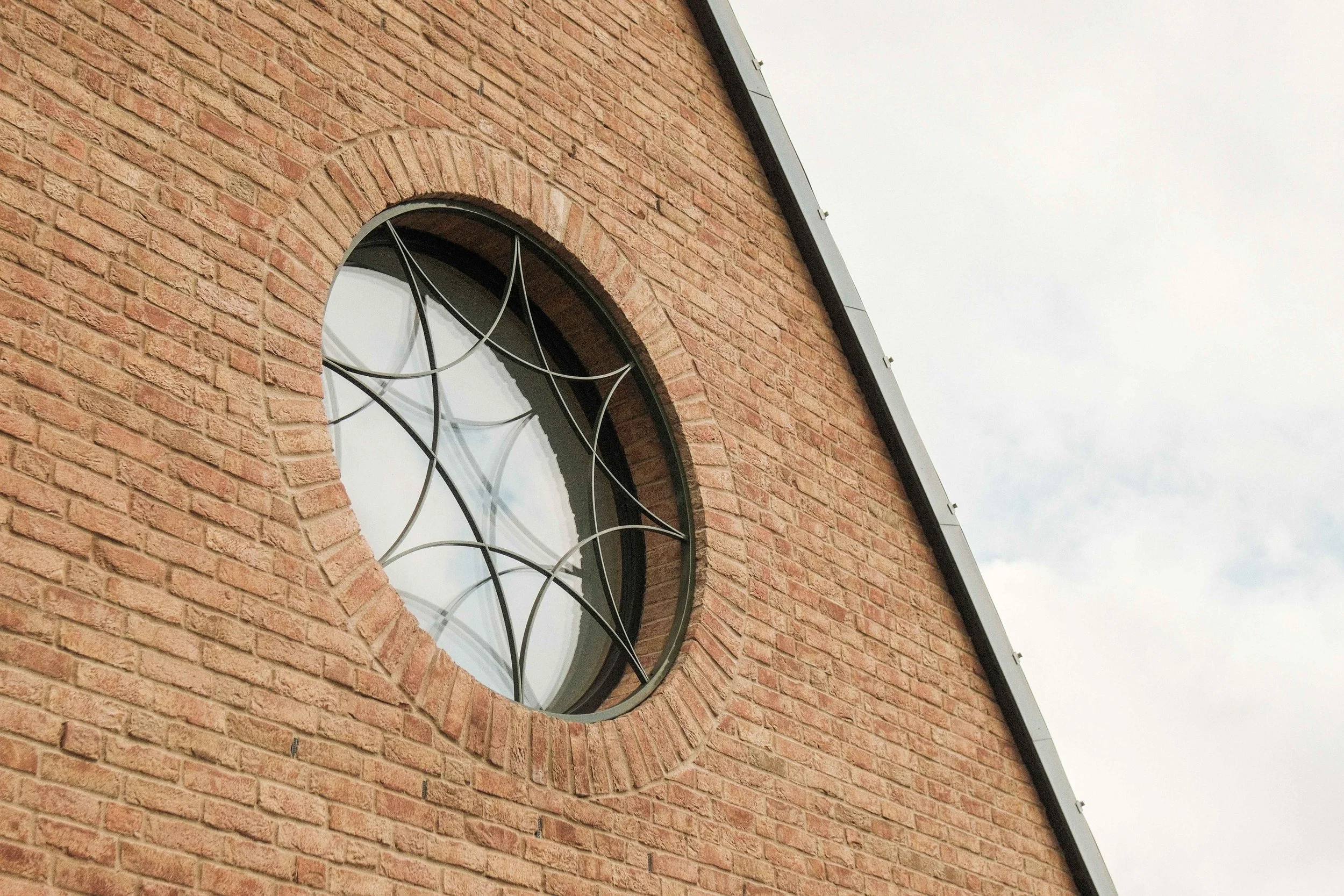 Close-up of a brick house with a circular window featuring a decorative metal grille, under a cloudy sky.