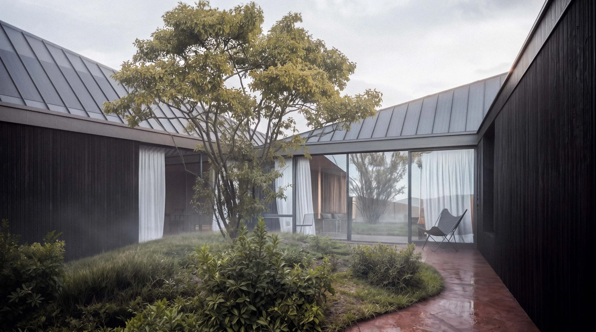 Modern house with black exterior walls and a metal roof, surrounded by greenery. A tree is in the courtyard with chairs near a glass wall.