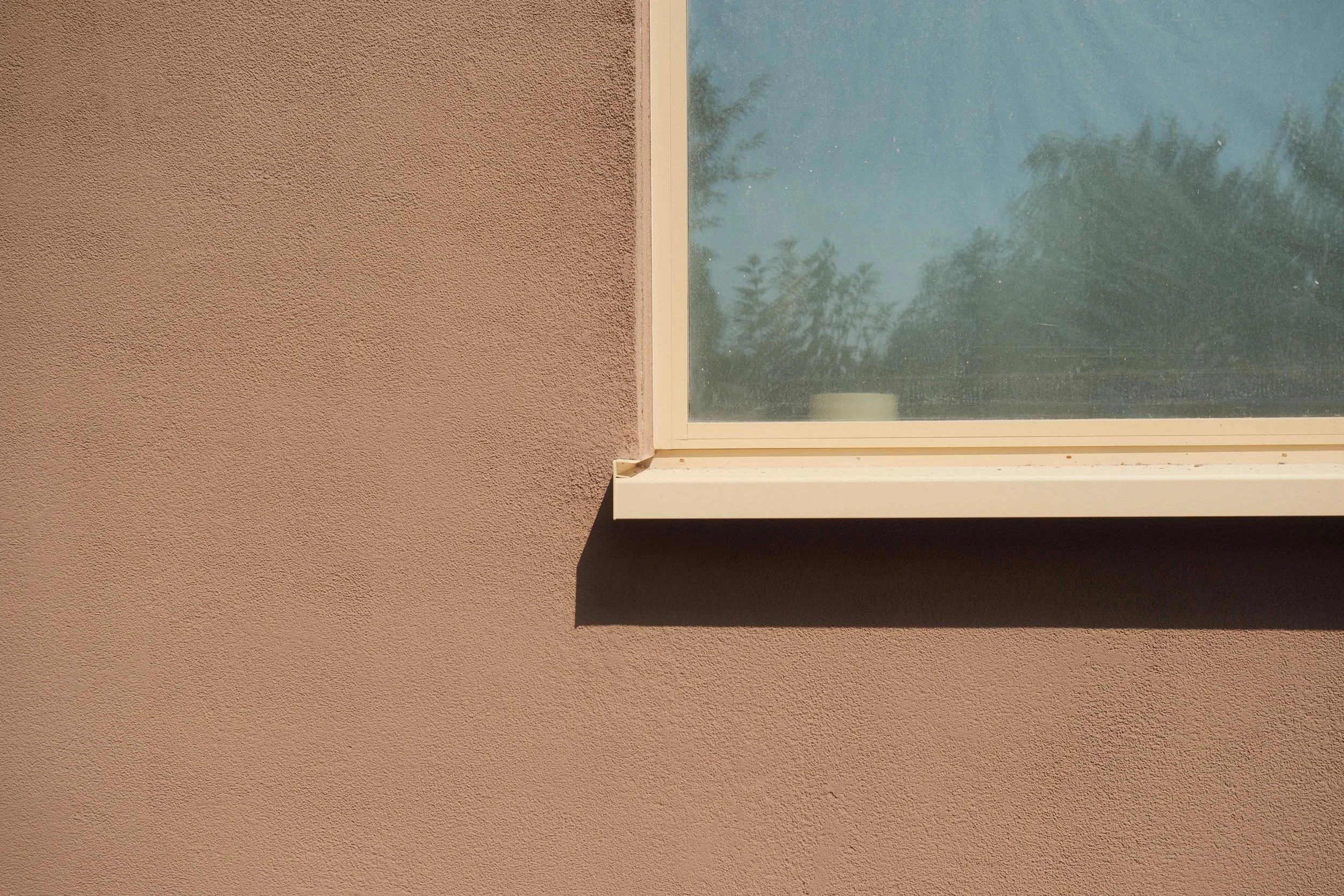 Close-up of a window set in a textured, beige wall. The window has a white frame and reflects trees and the sky outside.