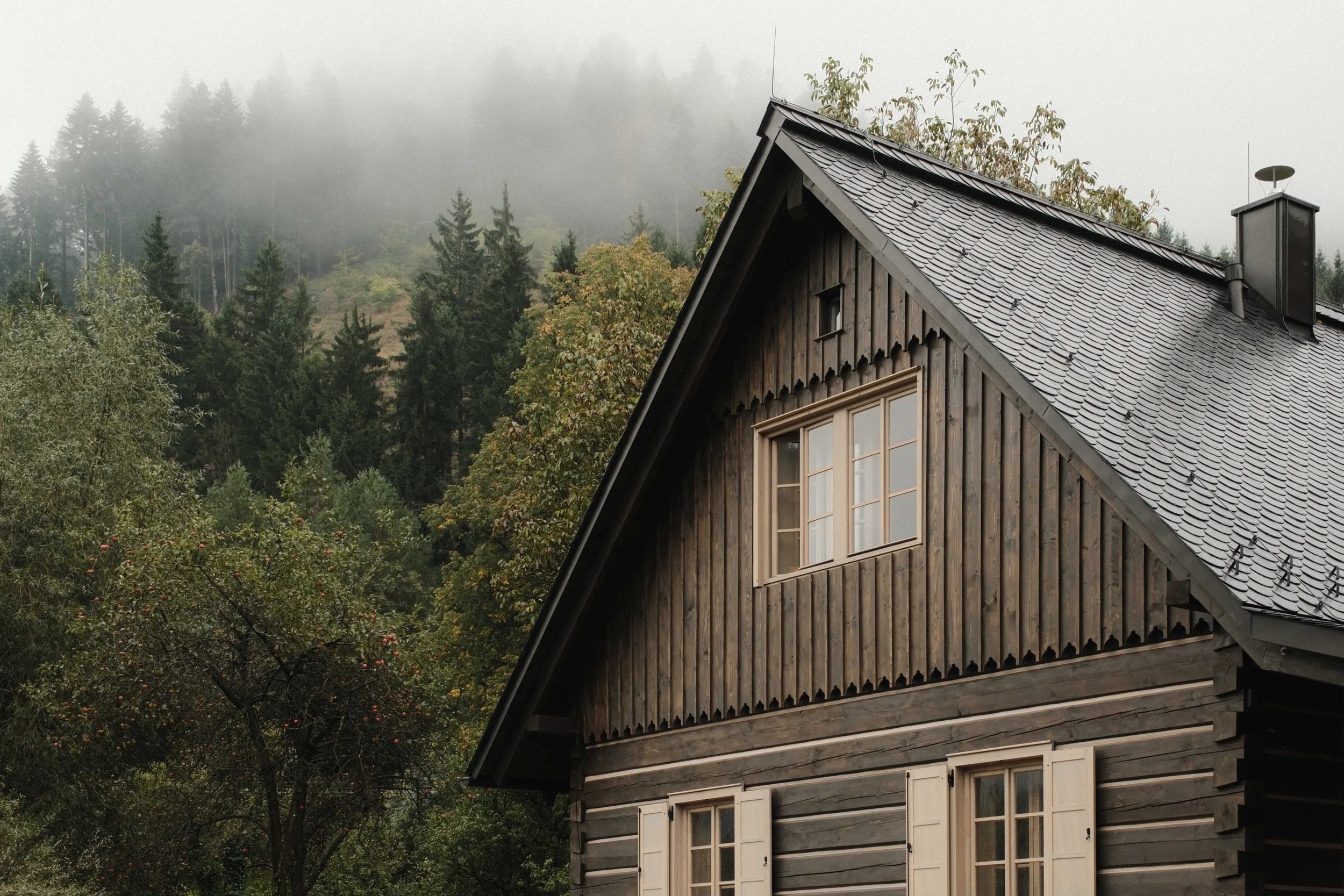 A wooden house with a steeply pitched roof covered in gray tiles, surrounded by green trees, with foggy mountains in the background.