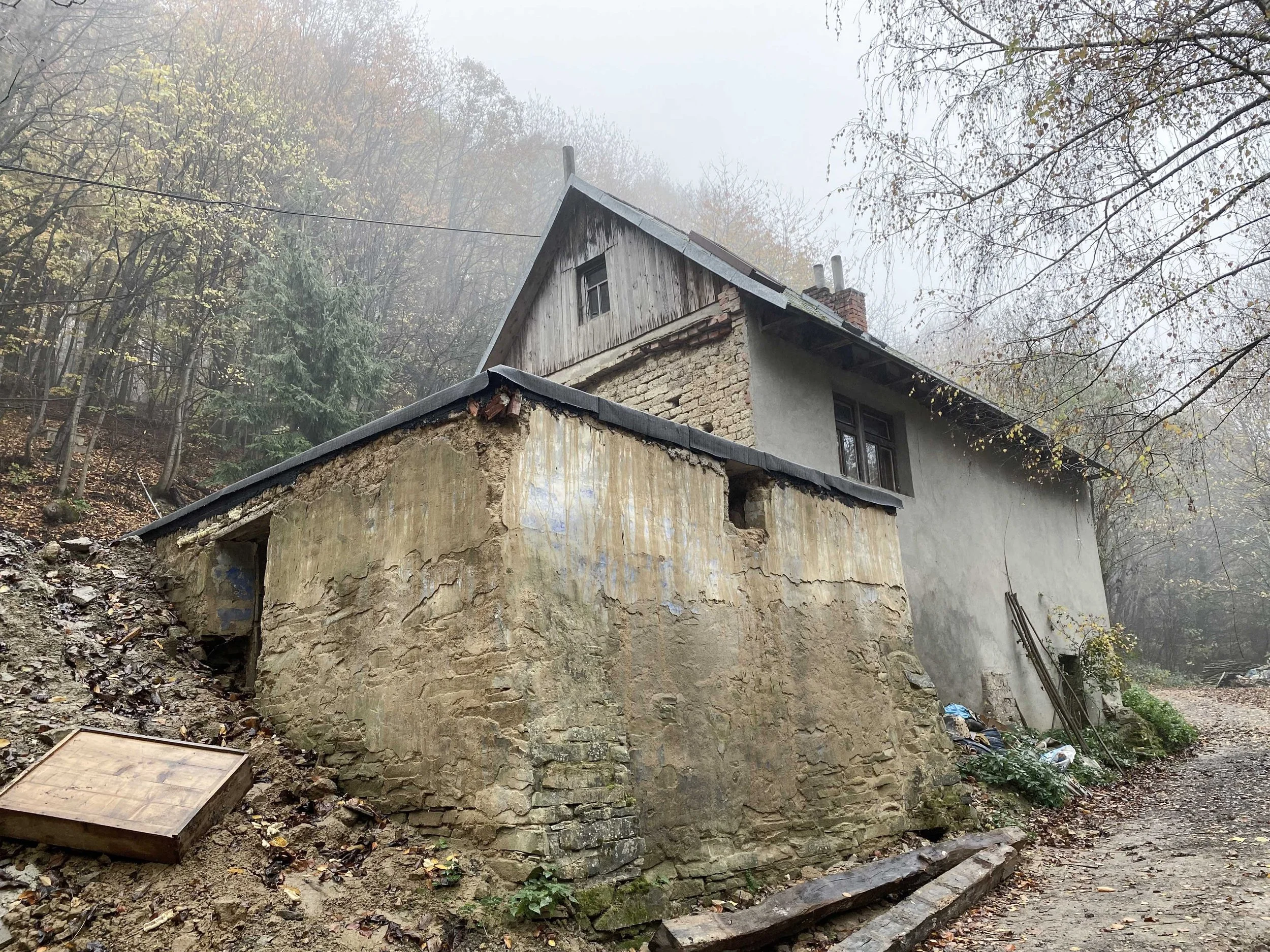 An old house built into a hill with a stone foundation, wooden upper walls, and a metal roof, surrounded by trees with sparse autumn foliage, in foggy weather.