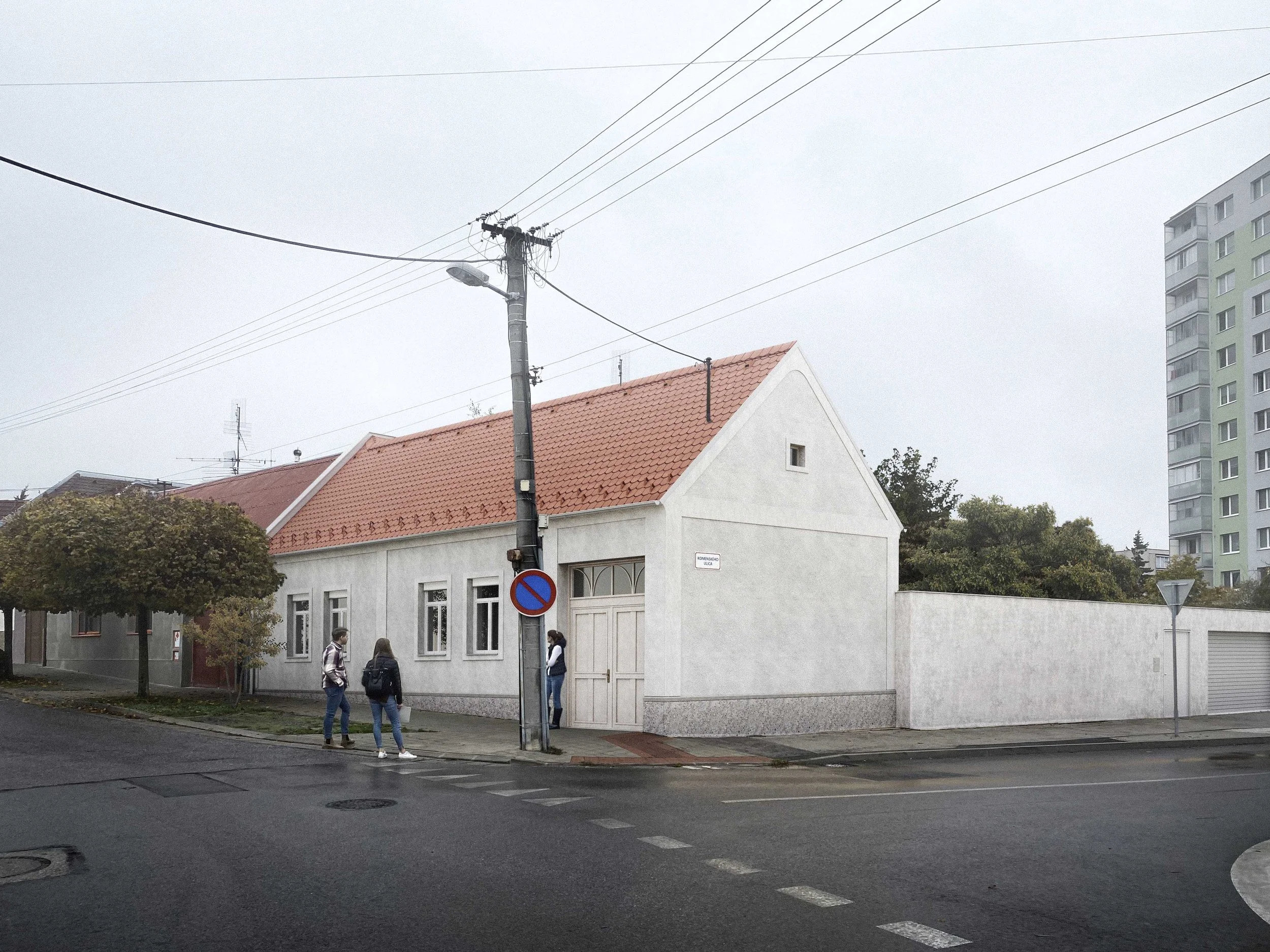 Street view showing a white building with a red tile roof, a tall utility pole with power lines, and some pedestrians walking on the sidewalk on a cloudy day.