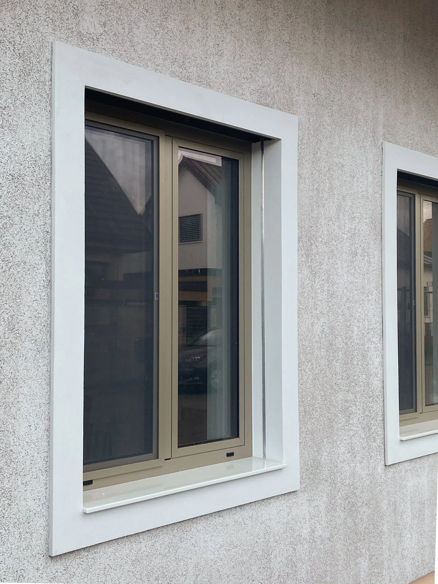 View of two window units with beige frames, installed on a wall with a stucco finish, surrounded by white trim and window sills.