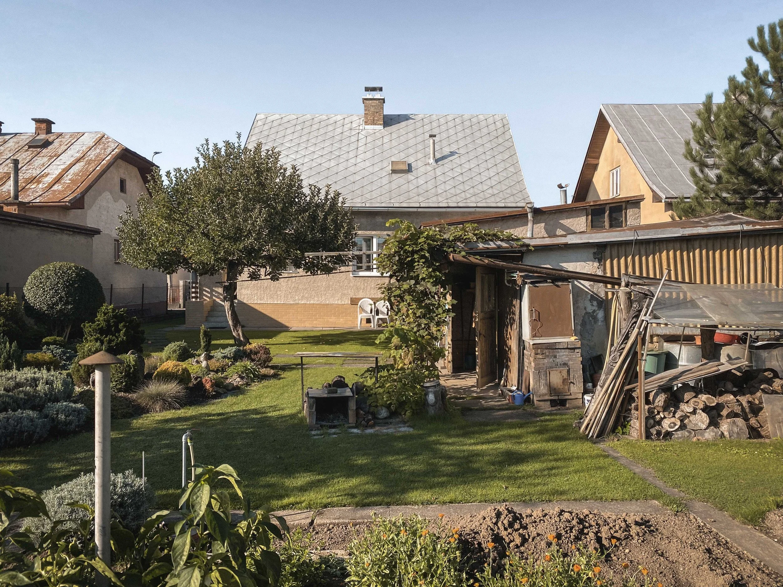 A backyard garden with trimmed bushes, a small tree, and a lawn with flower beds. There are houses in the background with metal and tile roofs, and various outdoor structures and gardening tools.