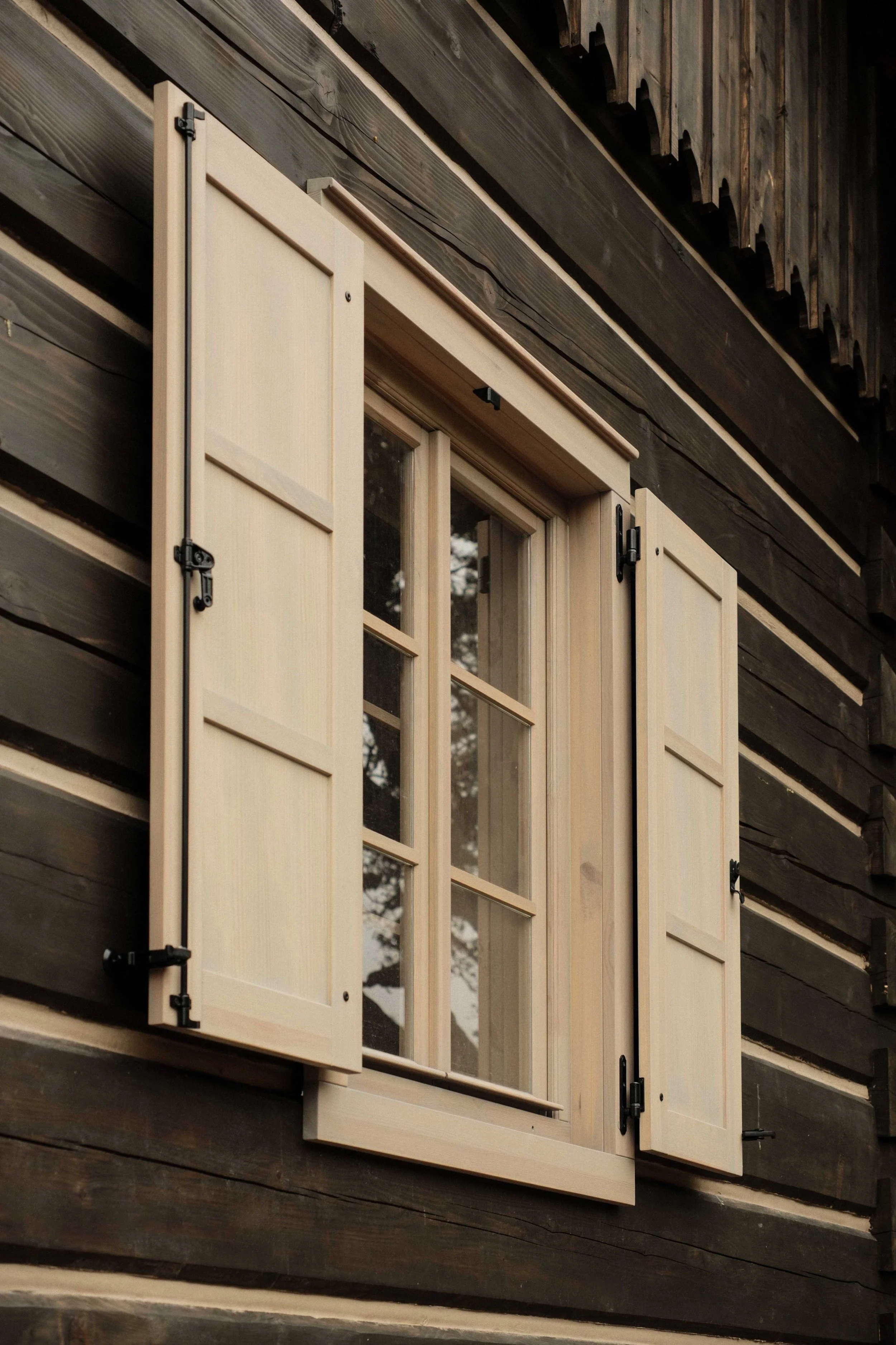 A window with wooden shutters mounted on a dark wooden house exterior, with the shutters open to reveal the window.