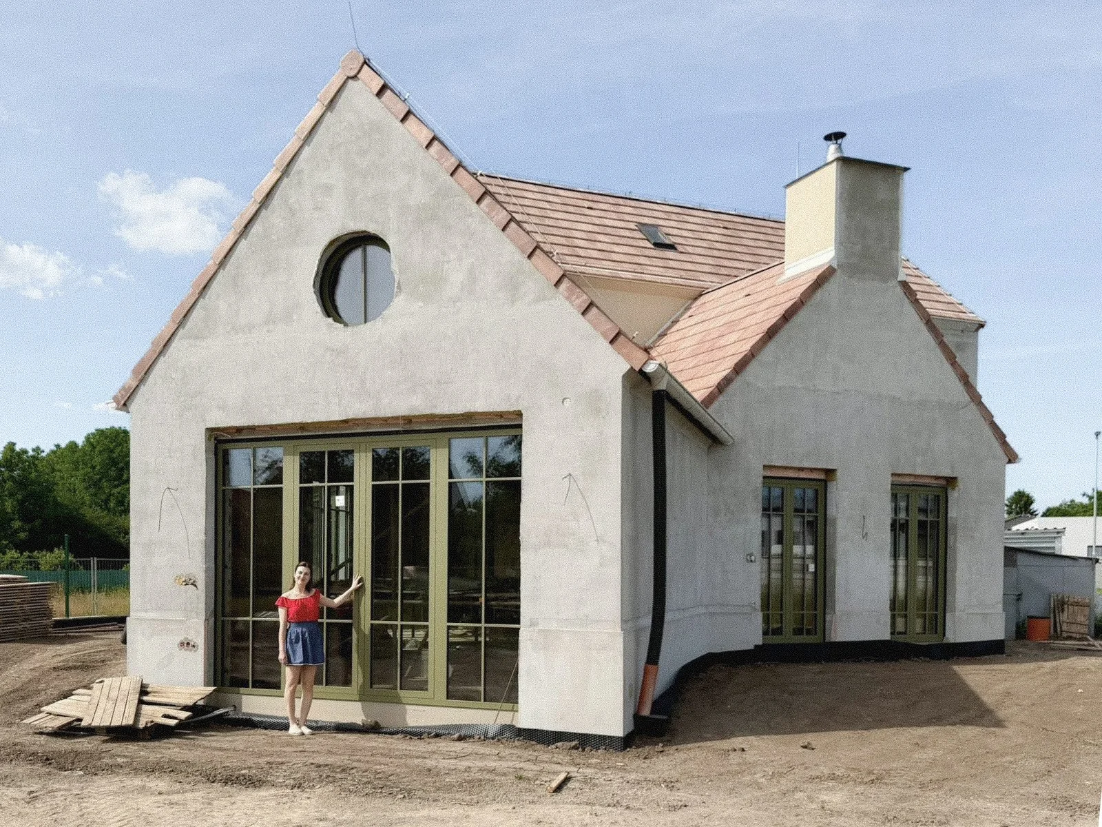 A house under construction with a woman standing in front of a large window, wearing a red shirt and a blue skirt, on a clear day with a blue sky.