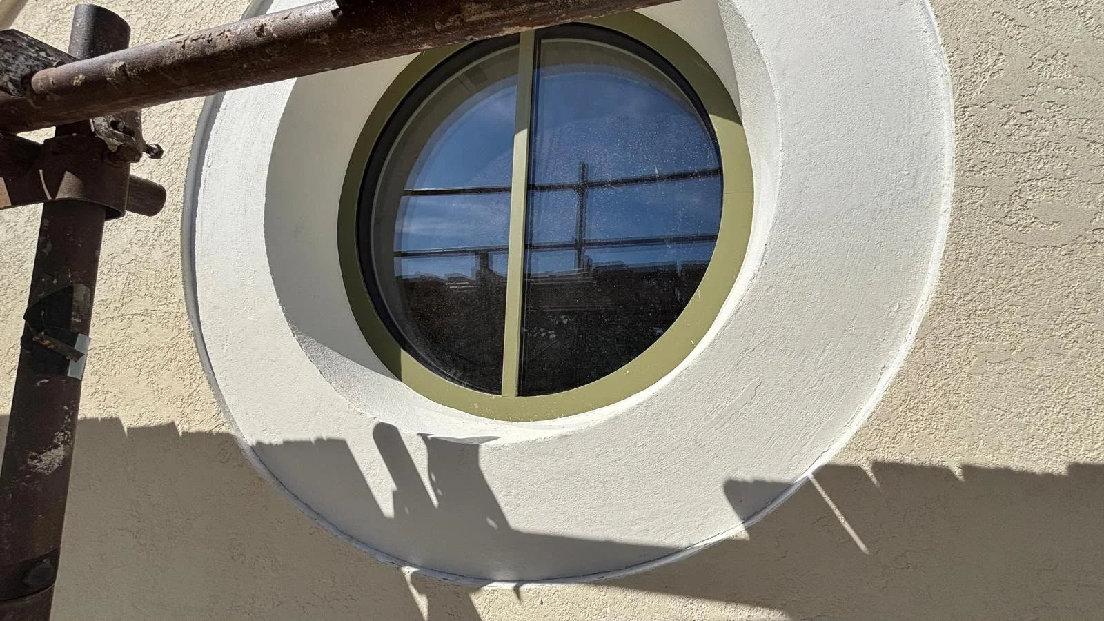 A round window with black metal framing and glass panes, set into a white textured wall. Shadows cast by scaffolding are visible on the wall.