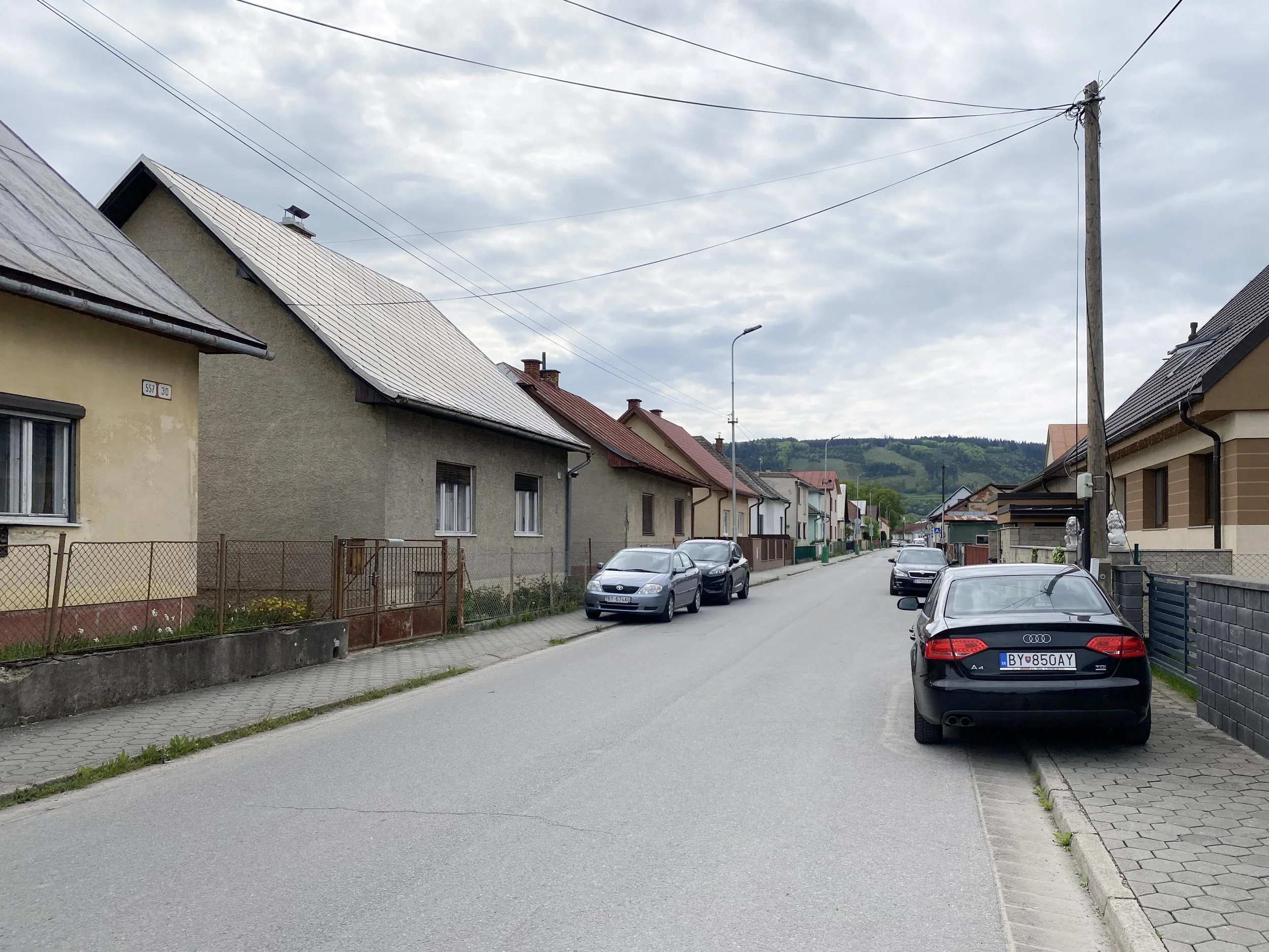 A residential street with parked cars, houses with sloped roofs, a sidewalk, and electrical wires in a small town or village setting, with green hills in the background.