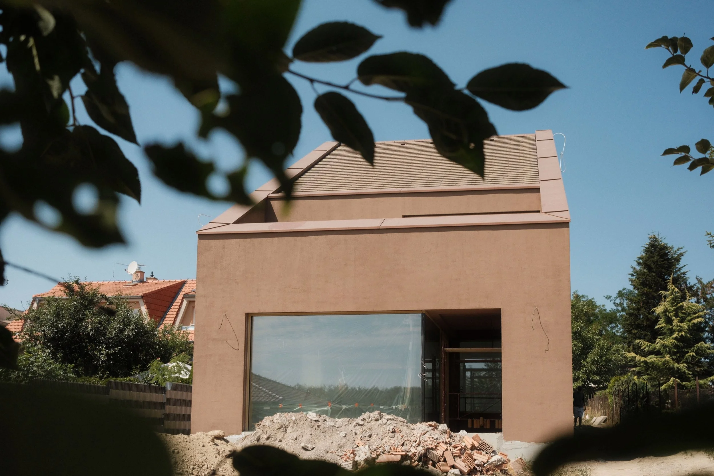 A modern building under construction with a large glass window, a beige exterior, and a section of the front wall open, revealing construction materials inside. The scene is framed by dark leaves in the foreground with a clear blue sky in the background.