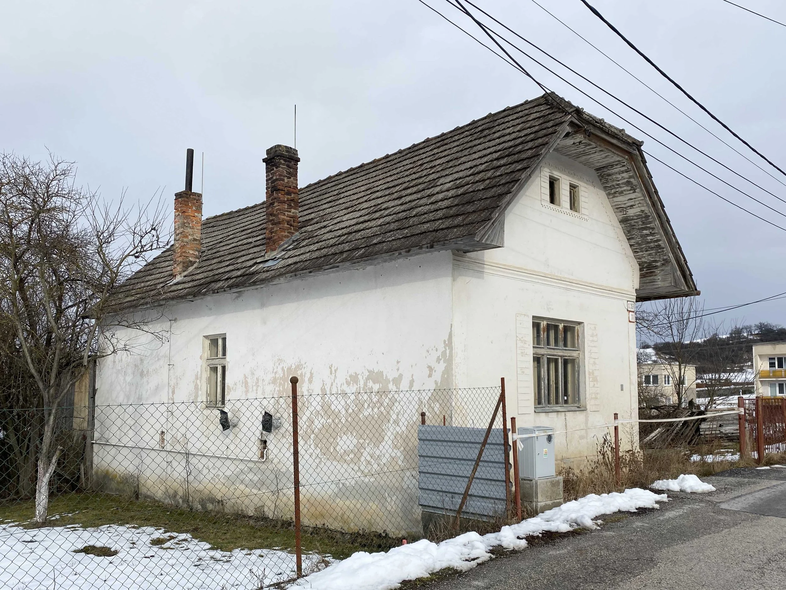 An old white house with a dark shingle roof, brick chimneys, and small windows. The house is behind a chain-link fence with some snow on the ground and leafless trees nearby. Overhead power lines are visible in the sky.