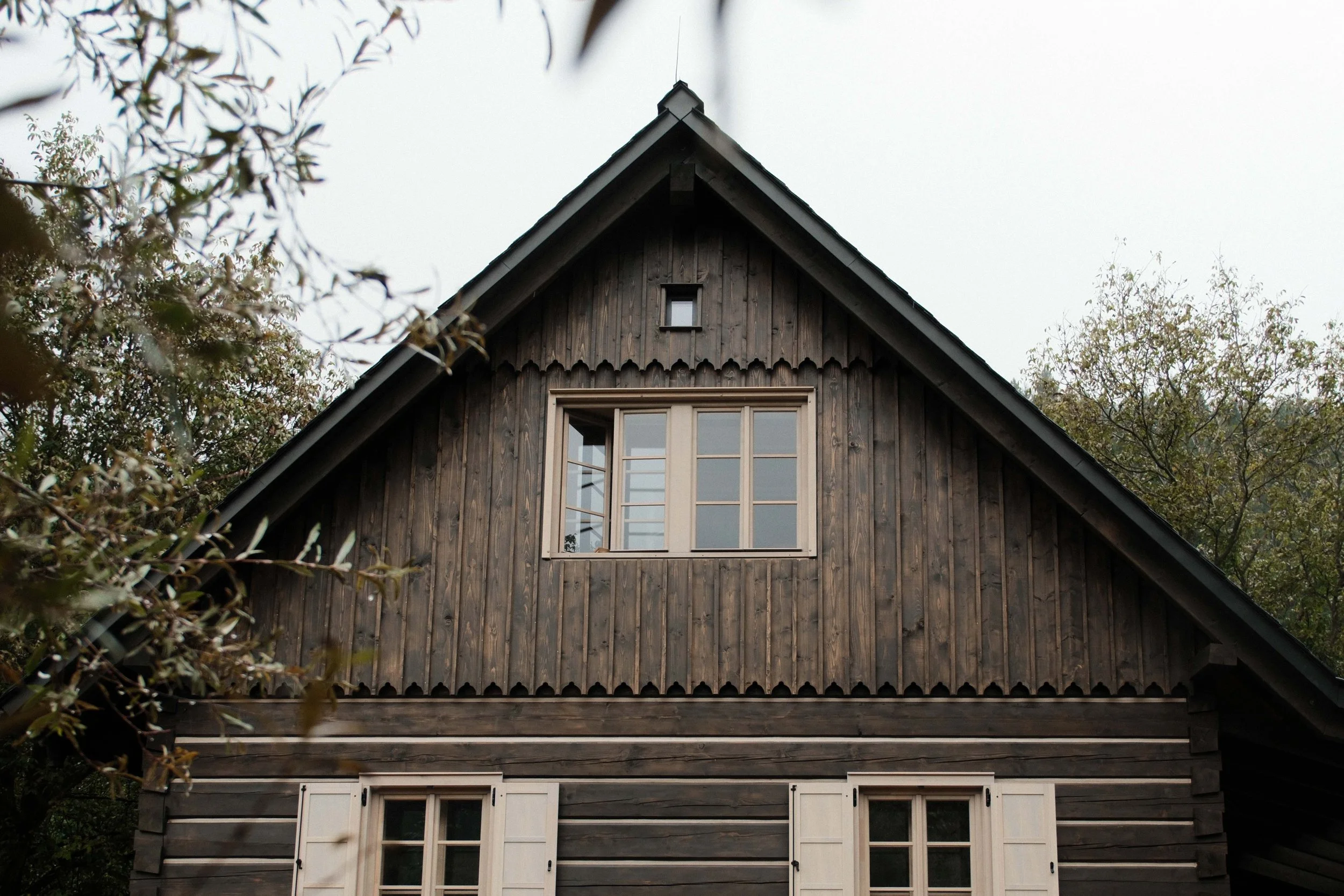 Rear view of a two-story wooden house with a steep gable roof, surrounded by trees and foliage.