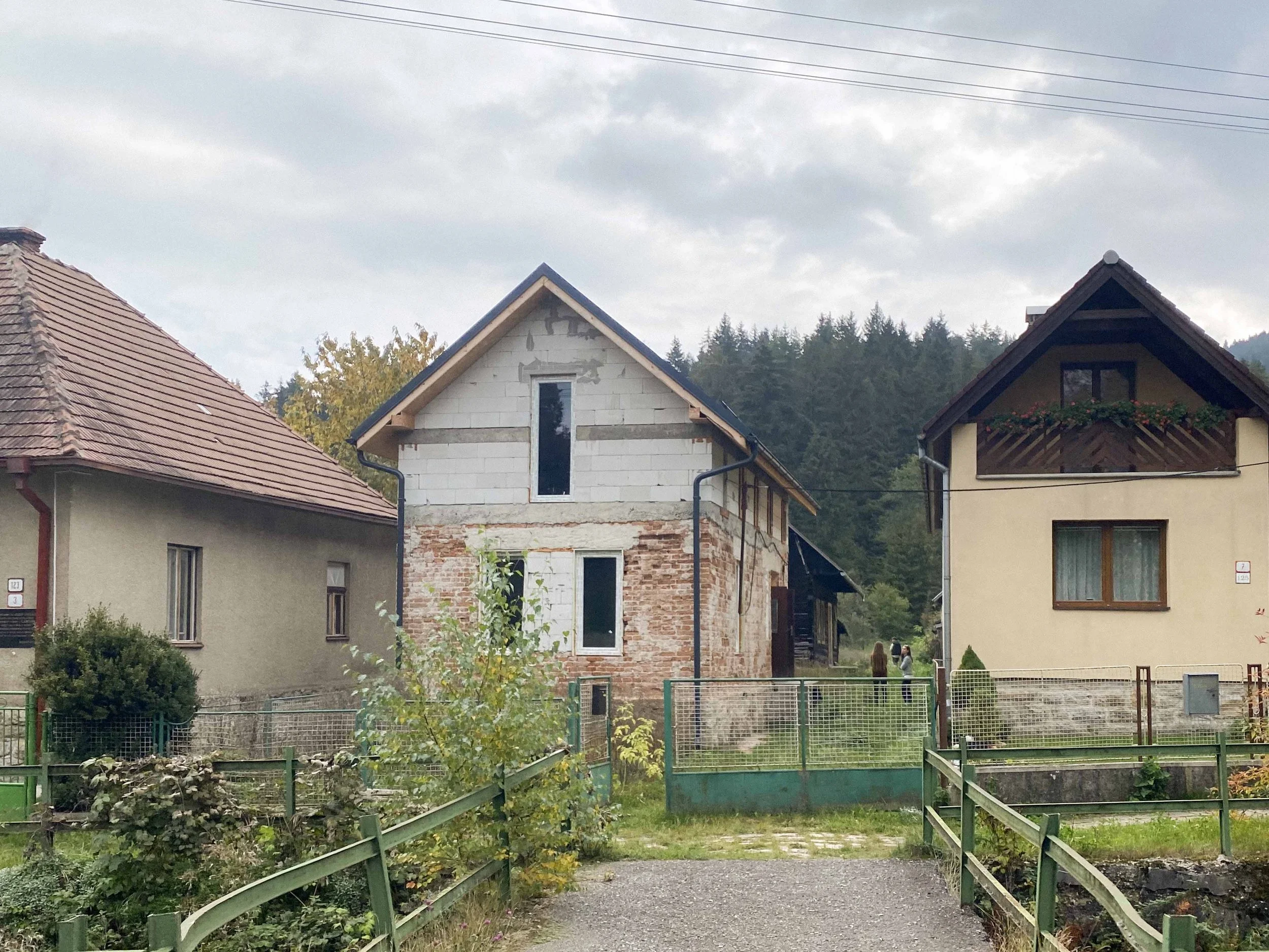 Neighborhood scene with unfinished house, neighboring houses, a garden, and a green fence in a hilly area.